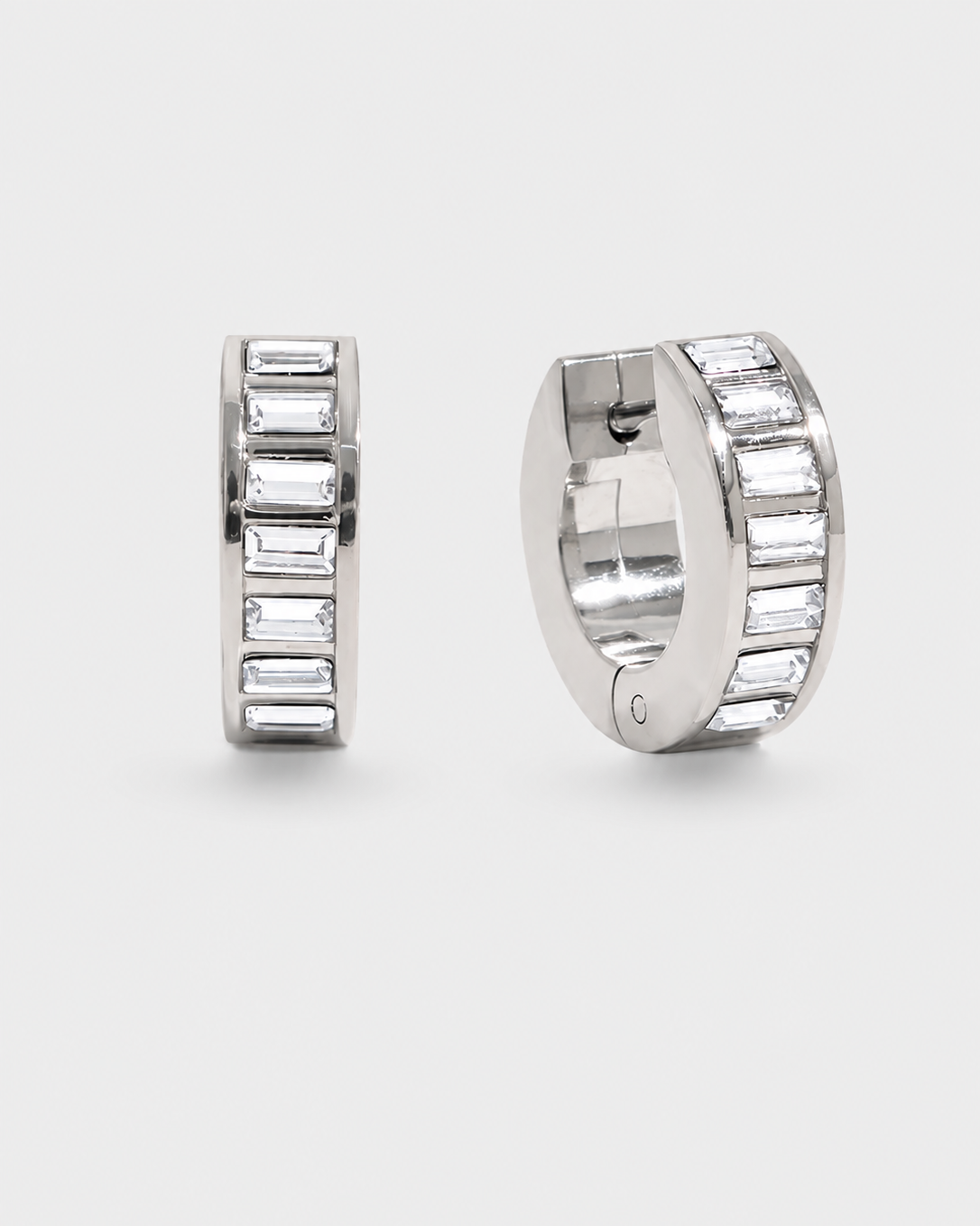 A pair of silver huggie hoop earrings embellished with baguette-cut clear crystals, shown from two angles on a light gray background.