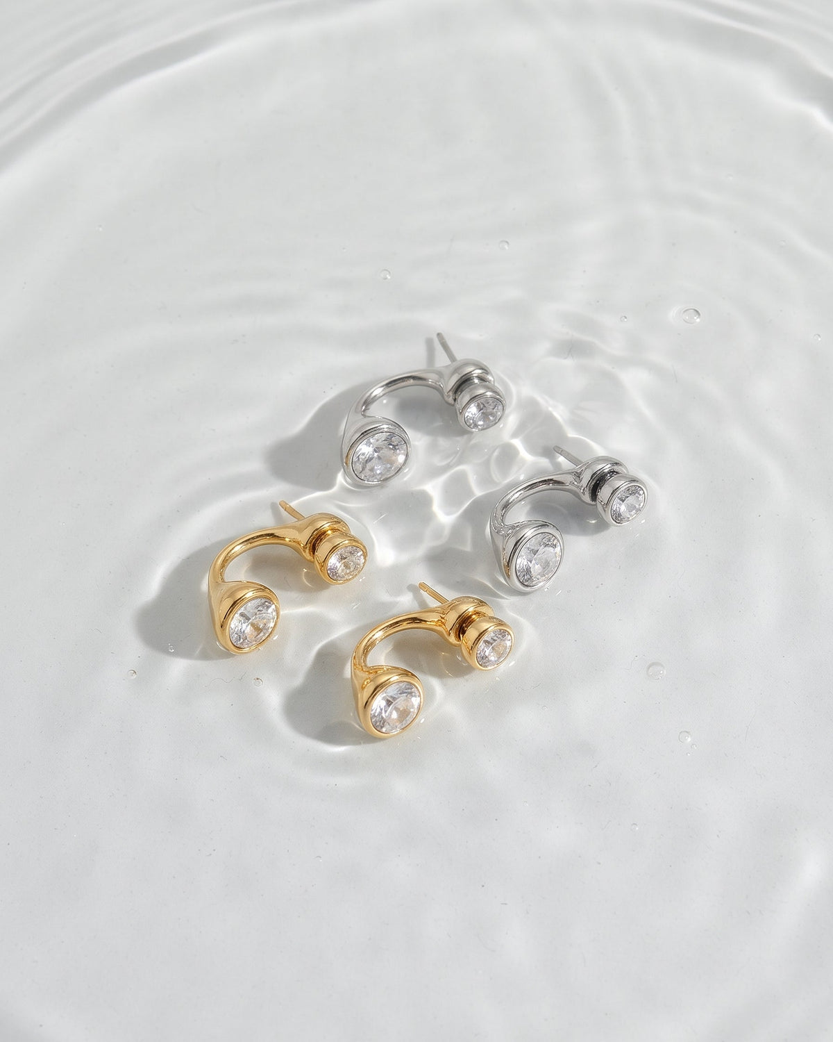 Four small hoop-style earrings with clear round stones, shown in two metal finishes: yellow gold and silver. They are arranged on a glossy white surface with soft water ripples around them.