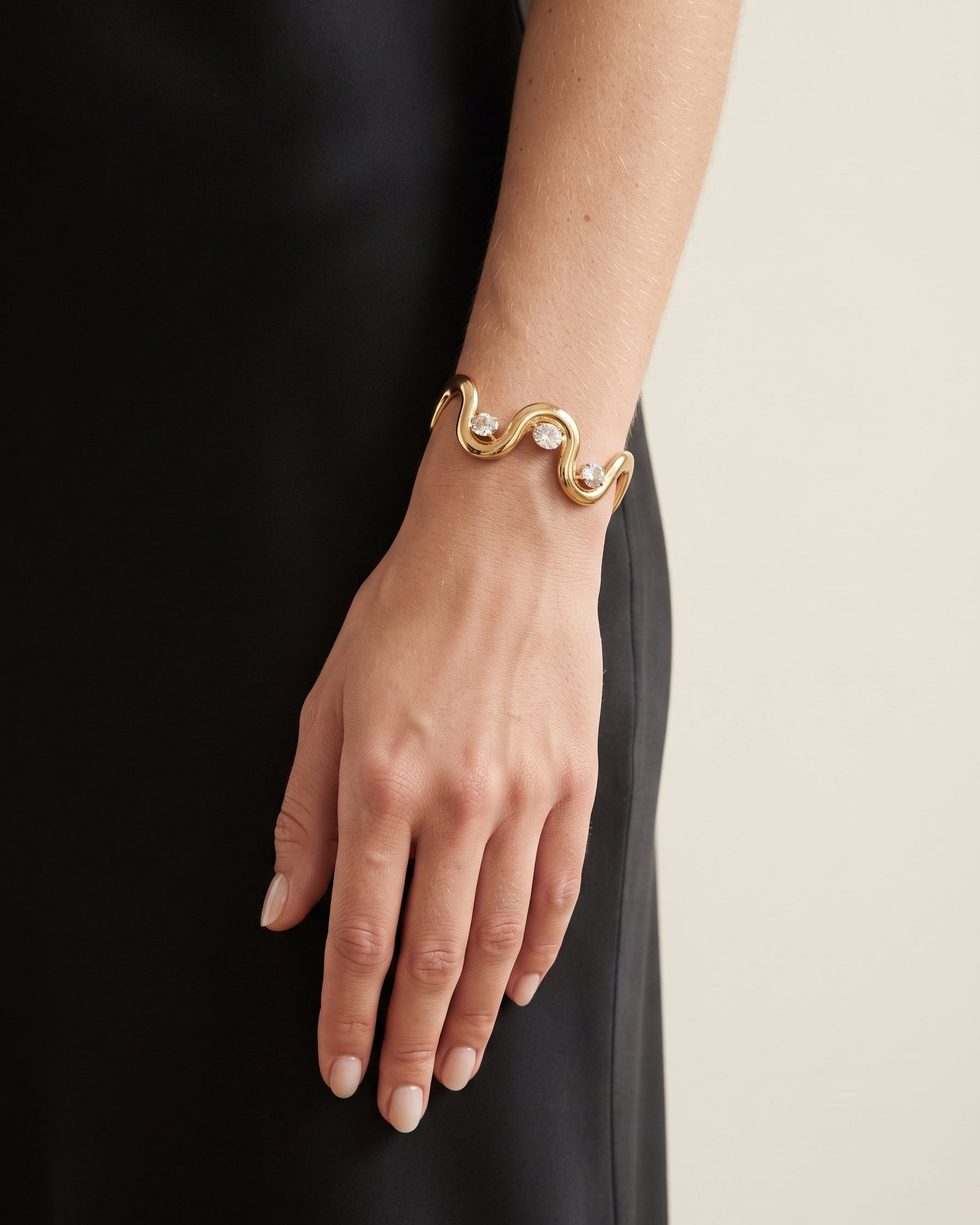 A gold wavy cuff bracelet with small clear stones, worn on a model's wrist against a black dress and a light beige background.
