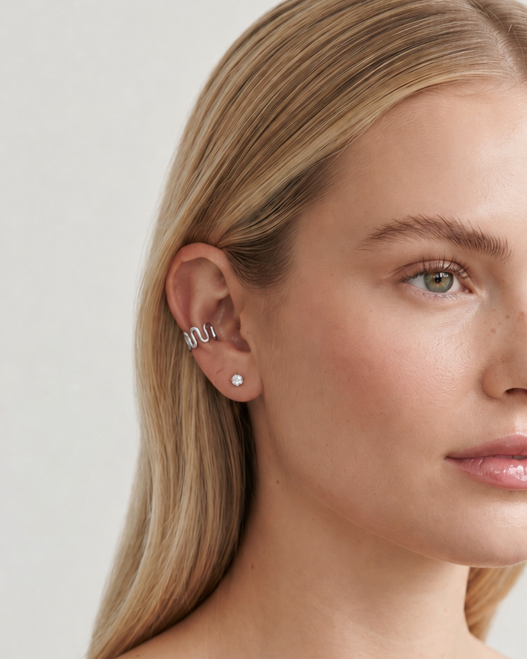 A close-up of a blonde woman wearing a stack of silver ear cuffs on her ear, paired with a black off-the-shoulder top against a light gray background.