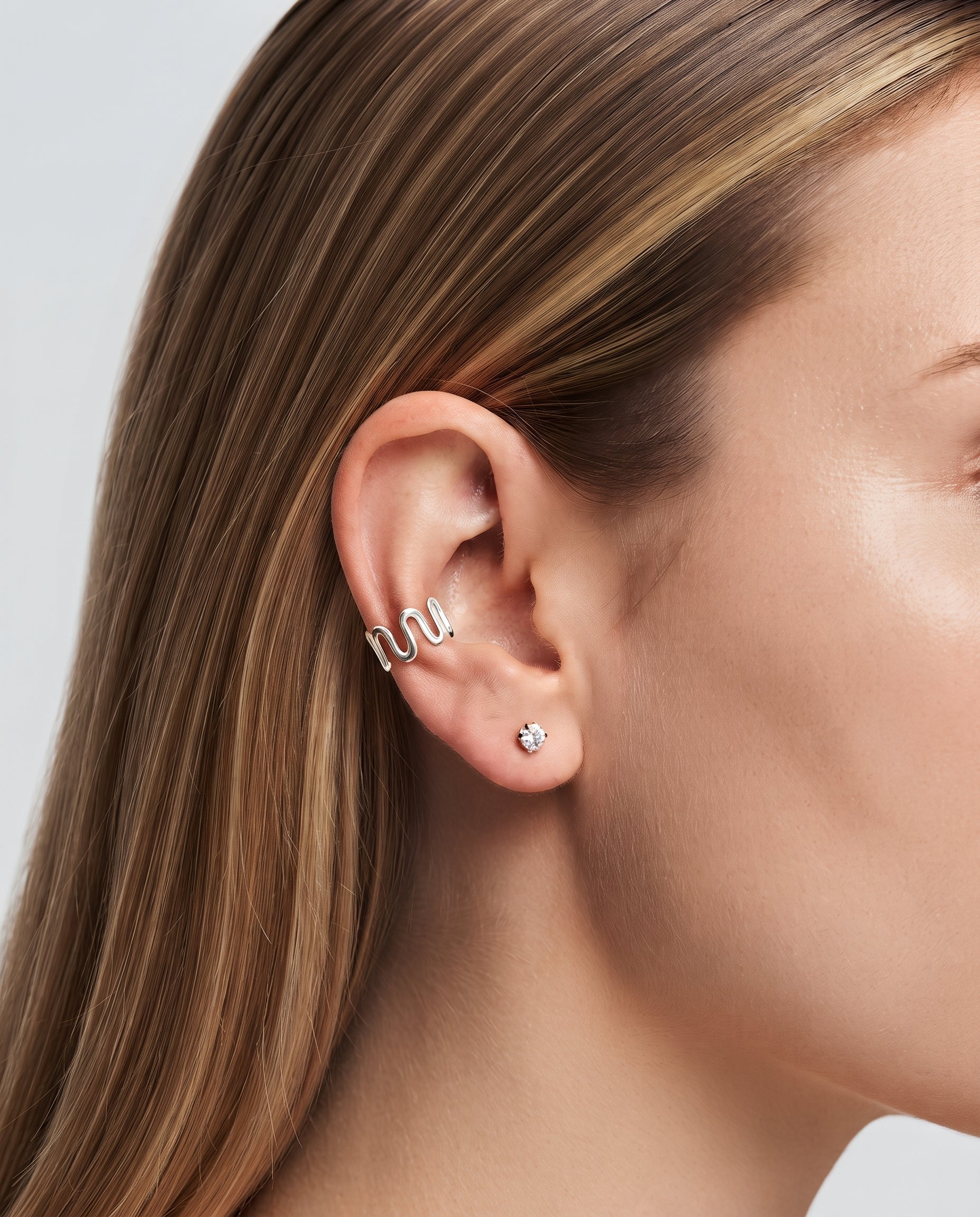 A close-up of a woman's ear showcasing a silver wire ear cuff with a wavy design and a small silver stud earring with a clear gemstone.
