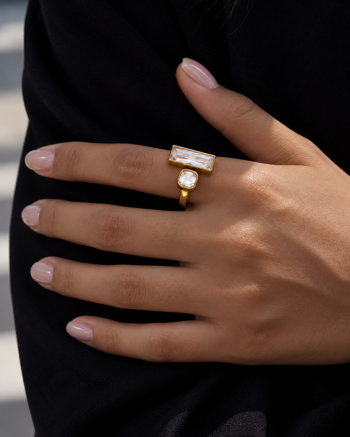 A close-up of a hand wearing two gold rings with clear stones, one rectangular and one oval, against a black sleeve.