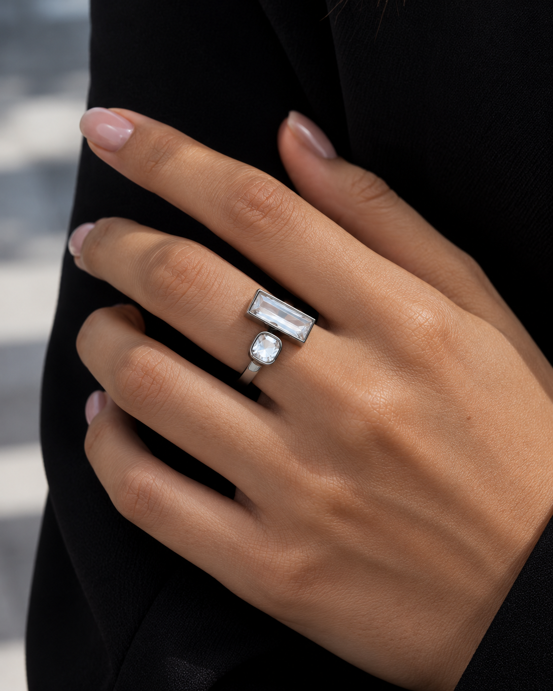 A close-up of a hand wearing a silver ring with a rectangular baguette-cut clear gemstone and a small round clear gemstone, set against a dark black sleeve.