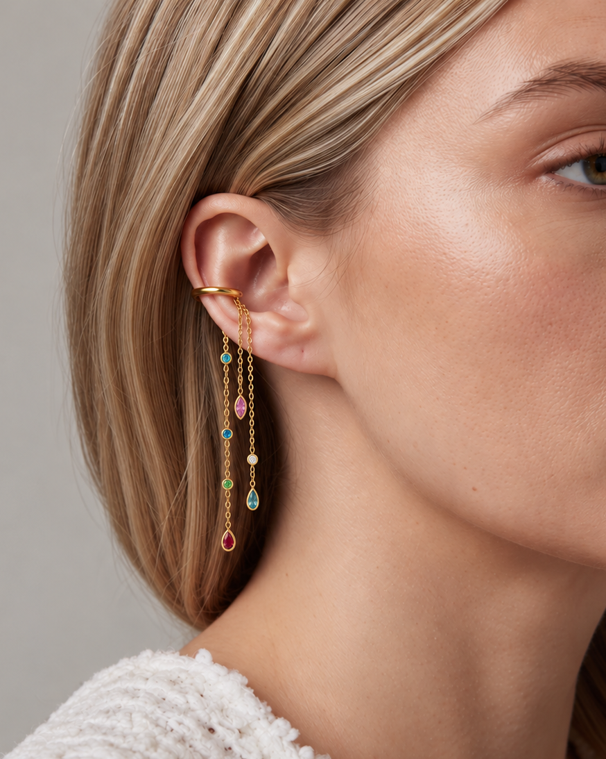 A close-up of a woman's ear wearing a gold huggie hoop earring with three dangling chains adorned with small gold beads and colorful teardrop gemstones in pink, green, and blue.