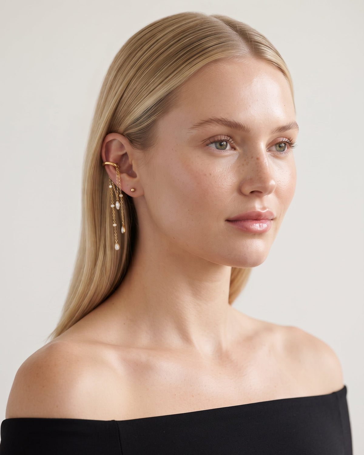A blonde woman wearing a delicate gold ear cuff with dangling bead chains, shown in a close-up portrait against a light neutral background.