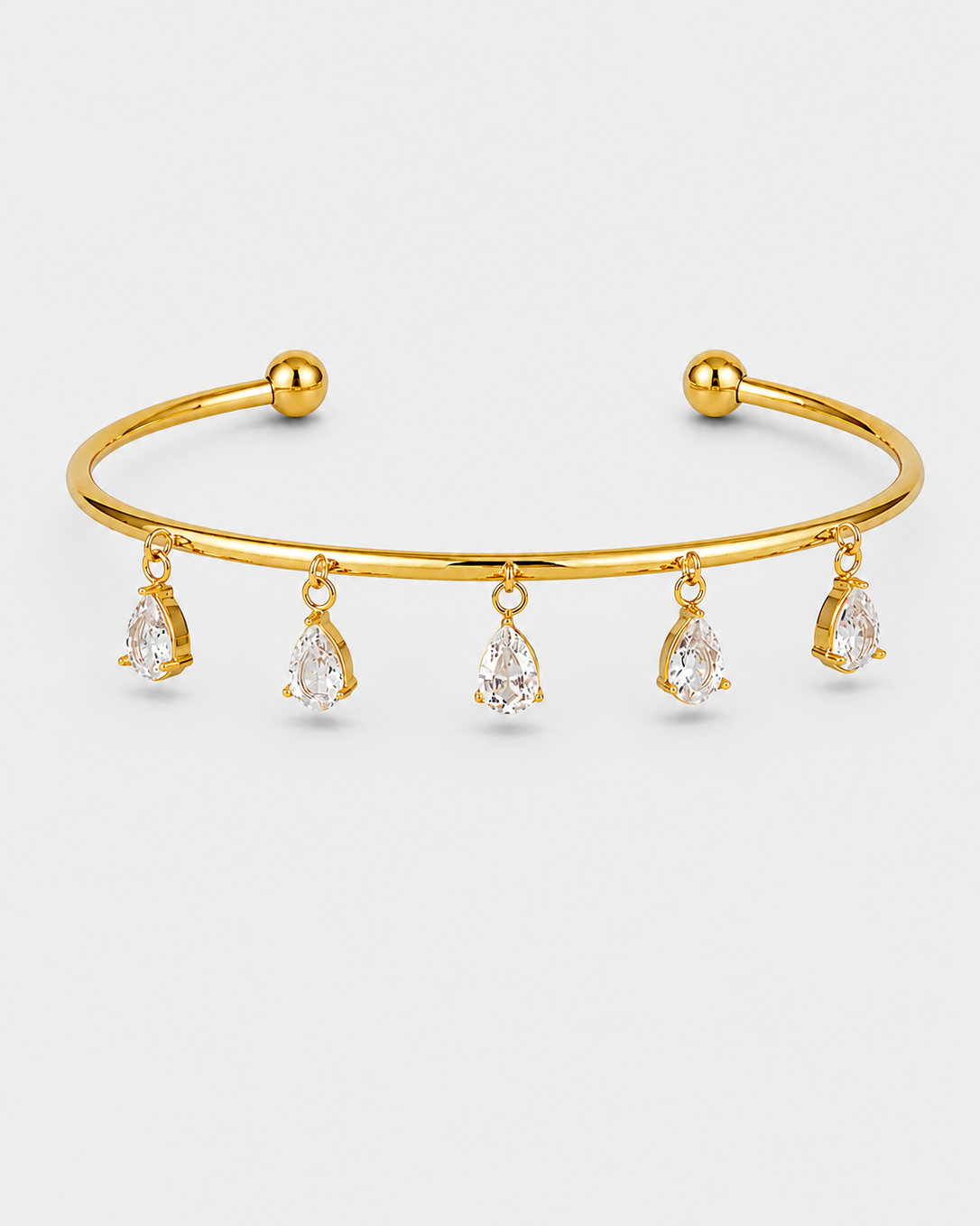A gold open cuff bracelet with five dangling pear-cut clear gemstones, shown on a light gray background.