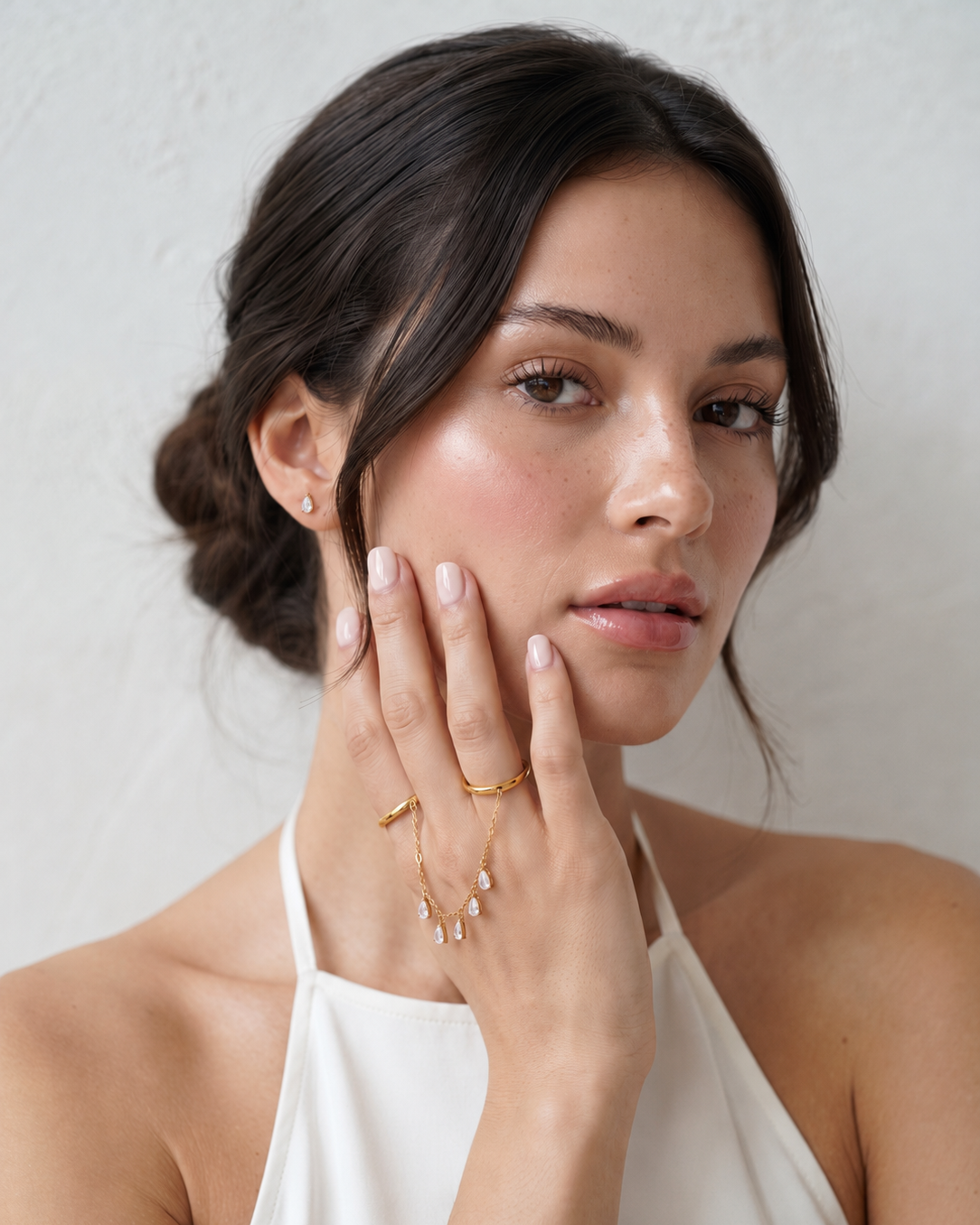 A close-up of a woman with dark hair in a low bun, wearing delicate gold rings and a gold chain necklace with small clear stones, paired with a white halter top against a light textured wall.