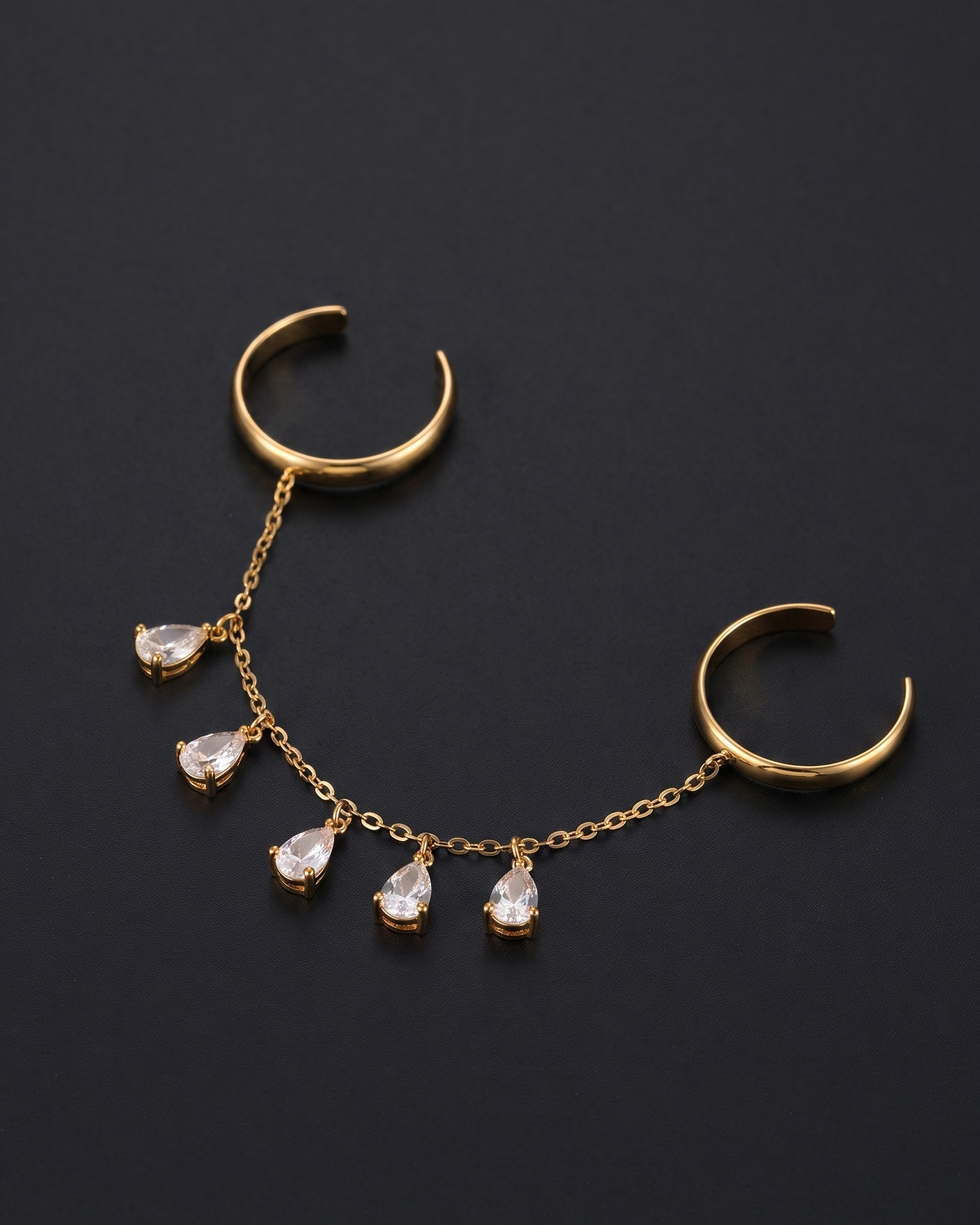 Gold-toned body jewelry featuring two curved hoop ends connected by a delicate chain with five dangling teardrop-shaped clear stones, displayed on a dark background.