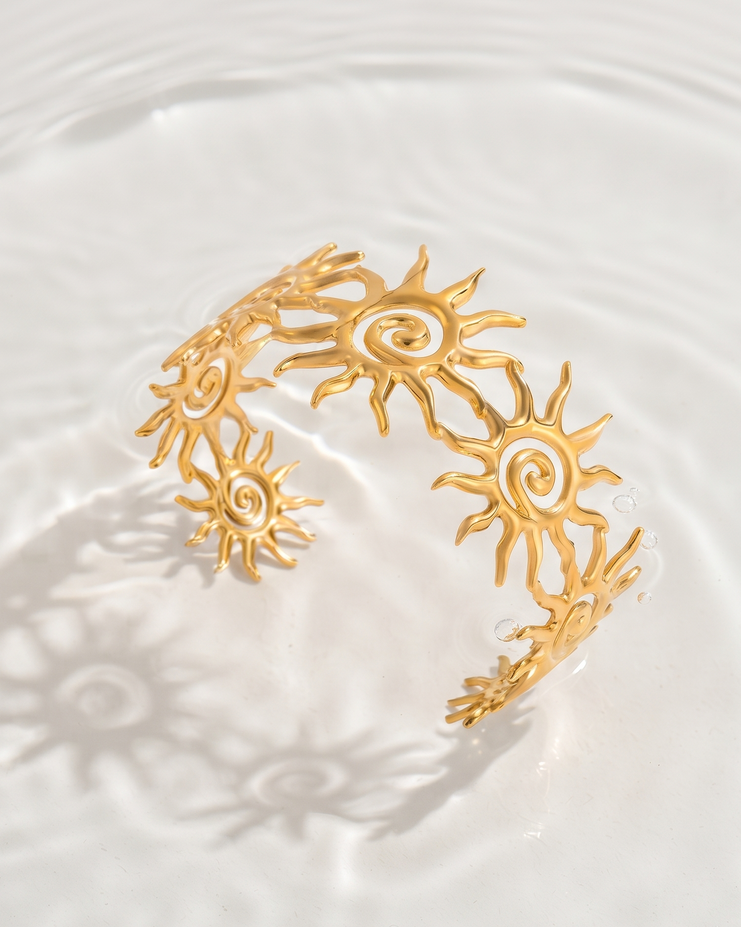 A gold-toned open cuff bracelet with a repeating sun motif, photographed floating in clear water with ripples and a sun shadow cast on the surface.