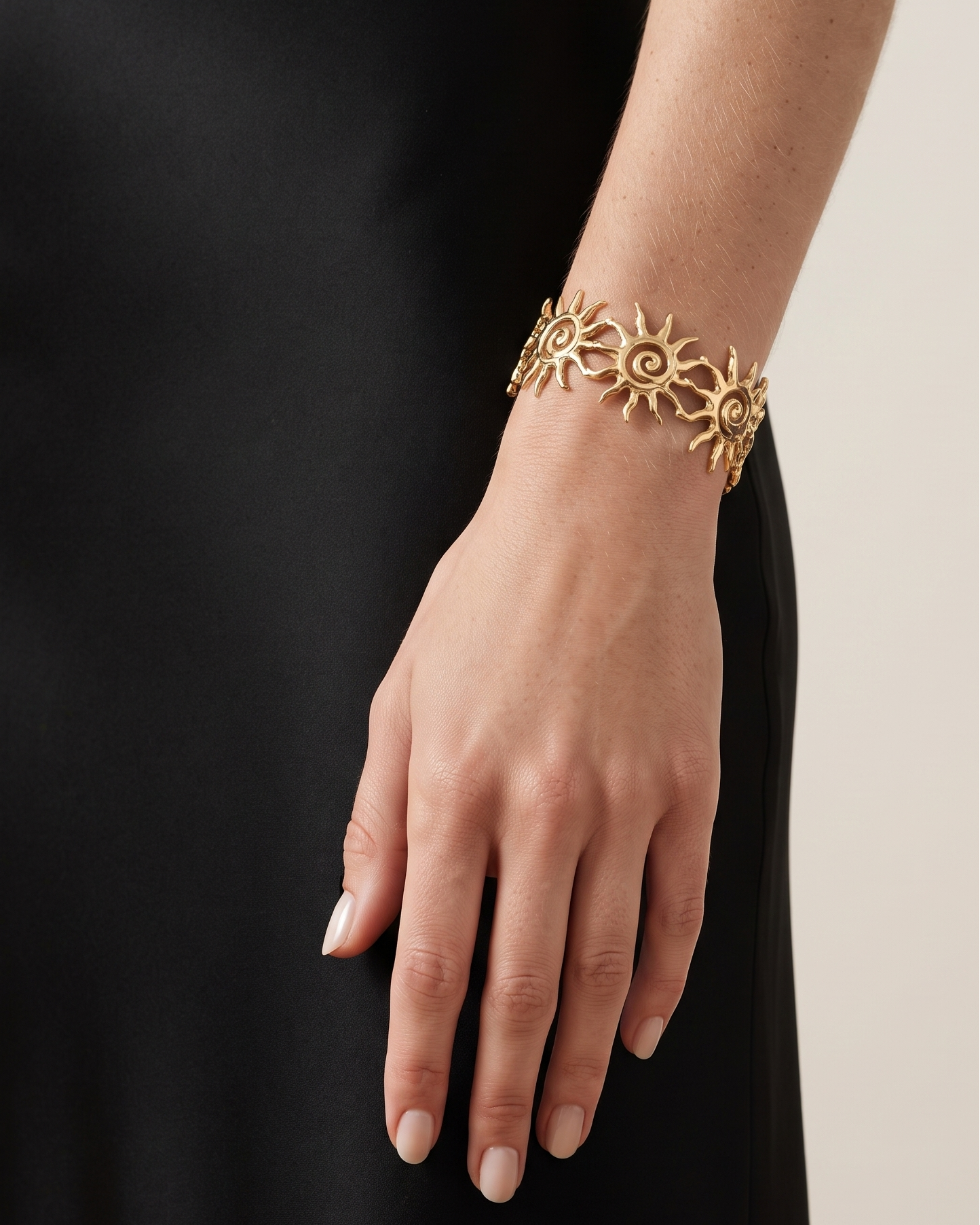 A gold-toned sunburst cuff bracelet worn on a model's wrist, paired with a black dress against a light beige background.