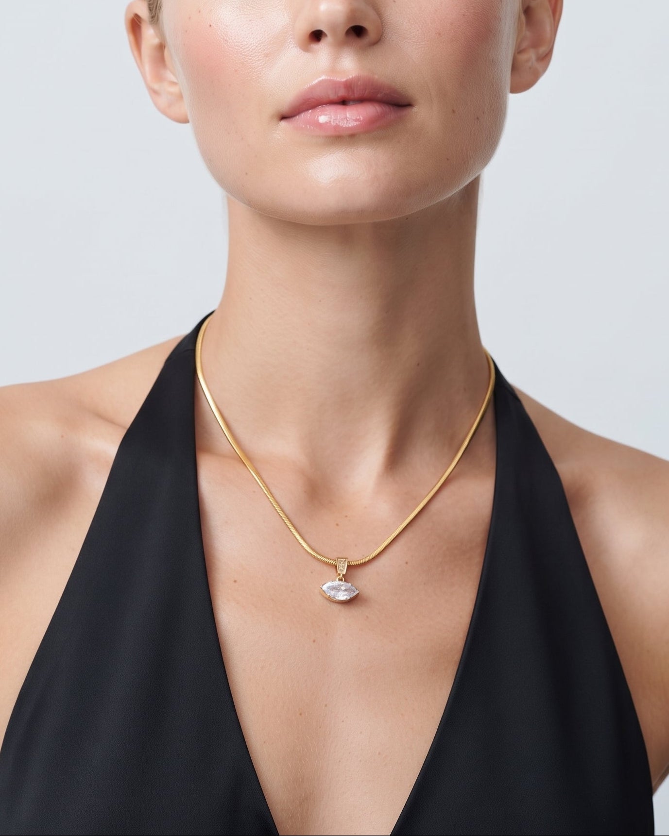 A blonde model wearing a delicate gold chain necklace with a small, clear, scalloped pendant, styled with a black halter top against a light gray background.