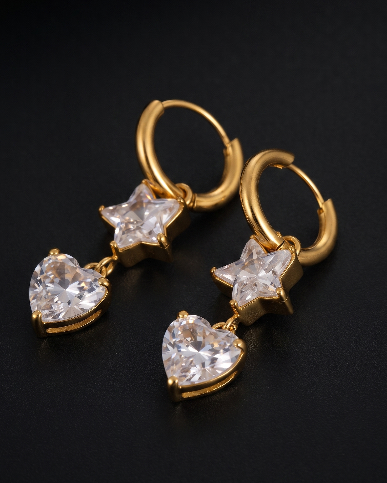 A pair of gold hoop earrings with dangling star and heart charms, featuring clear faceted gemstones, displayed on a dark black background.