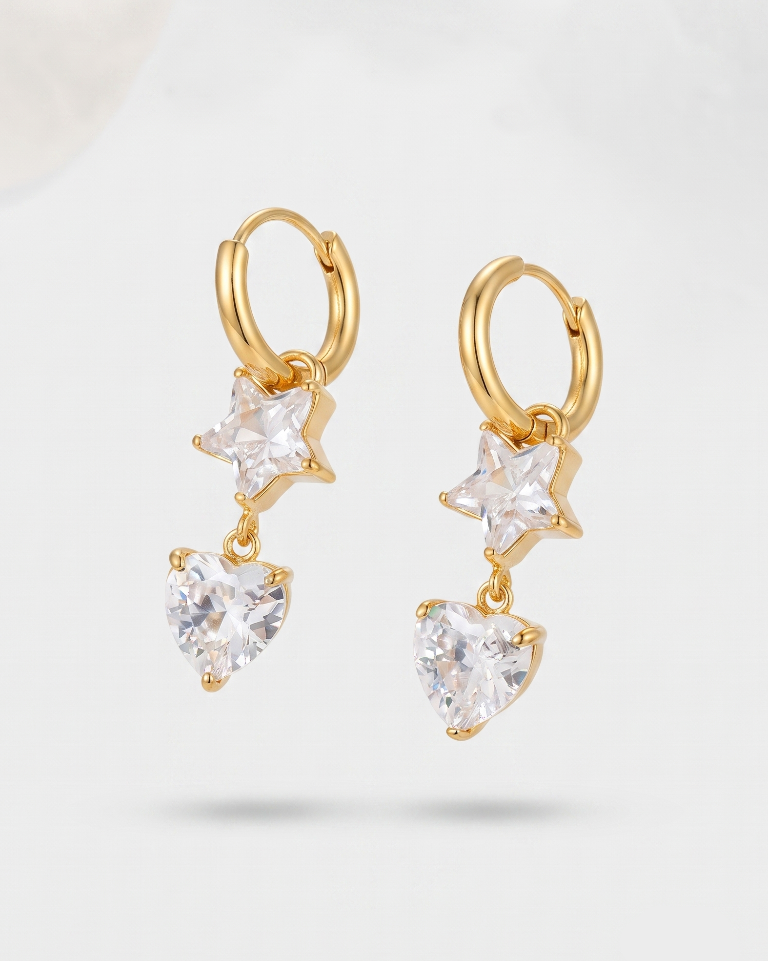A pair of gold huggie hoop earrings featuring a dangling star and heart design, set with clear cubic zirconia stones, shown against a light gray background.