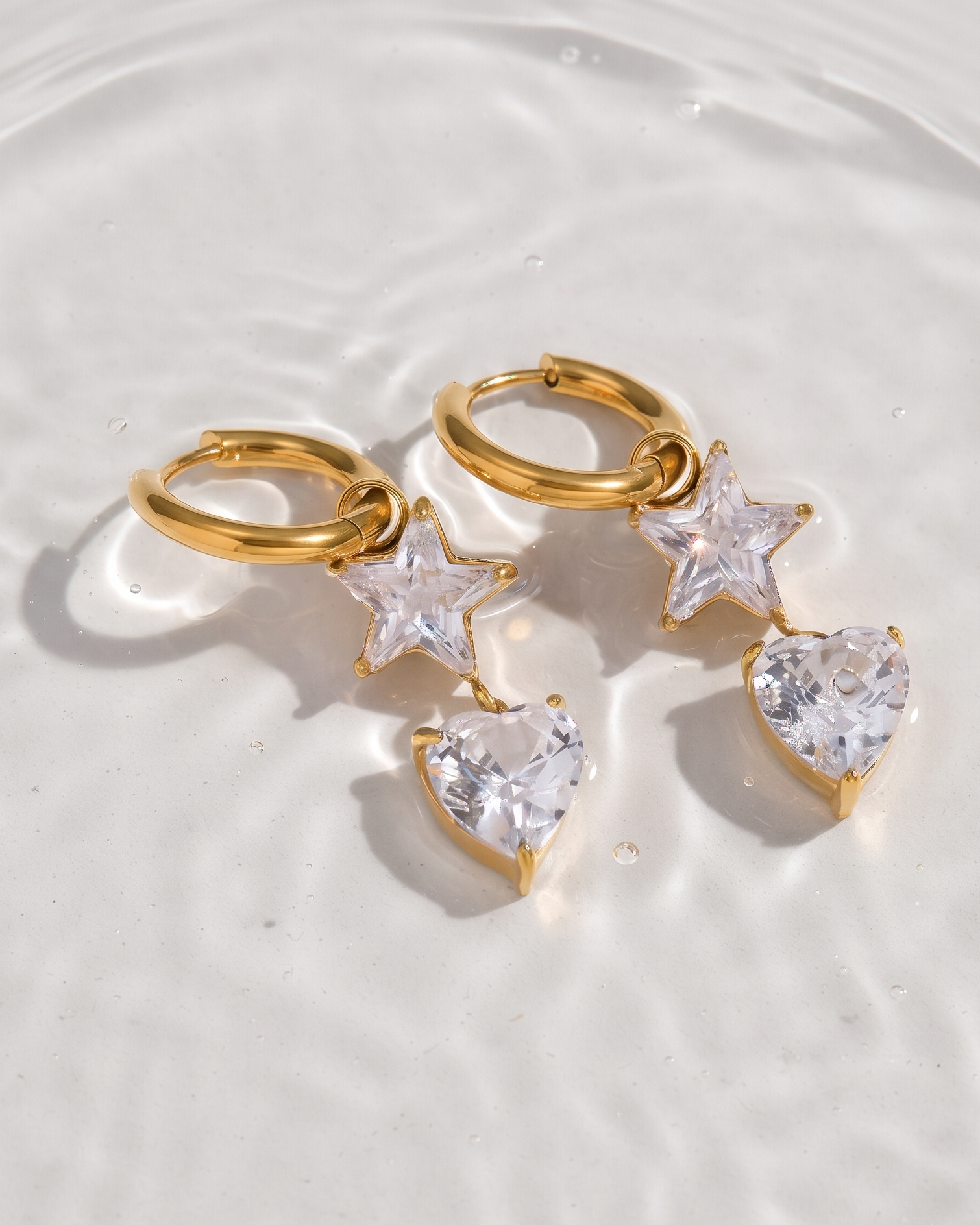 A pair of gold huggie hoop earrings with dangling star and heart charms, displayed on a white surface with water ripples.