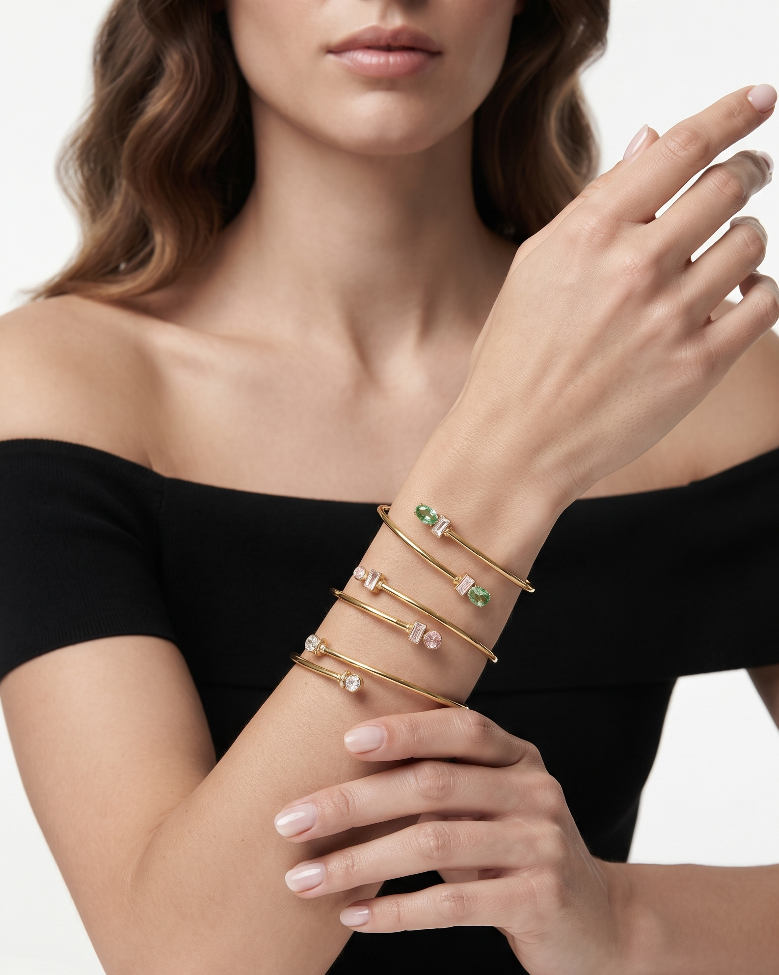 A close-up of a model wearing a stack of gold bangle bracelets with small green, pink, and clear gemstones on her wrist, paired with a black off-the-shoulder top against a clean white background.