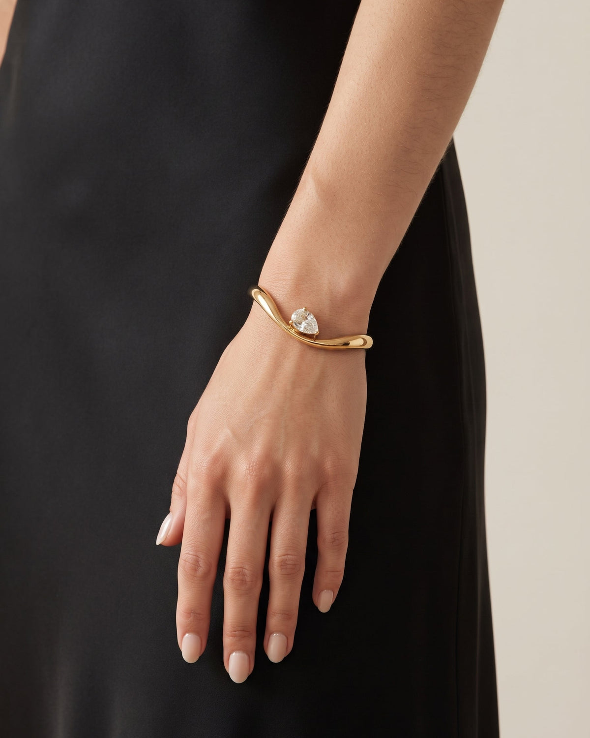 A gold curved cuff bracelet with a clear heart-shaped crystal centerpiece, shown worn on a model's wrist against a black dress and light beige background.