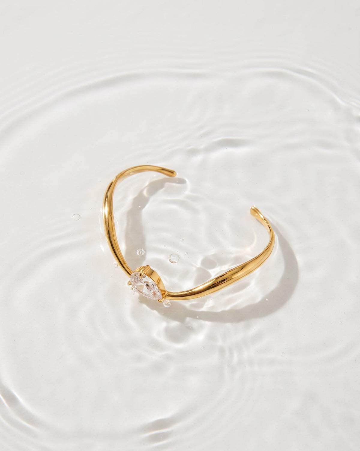 A gold open cuff bracelet with a clear teardrop gemstone, resting on a surface of rippling water.