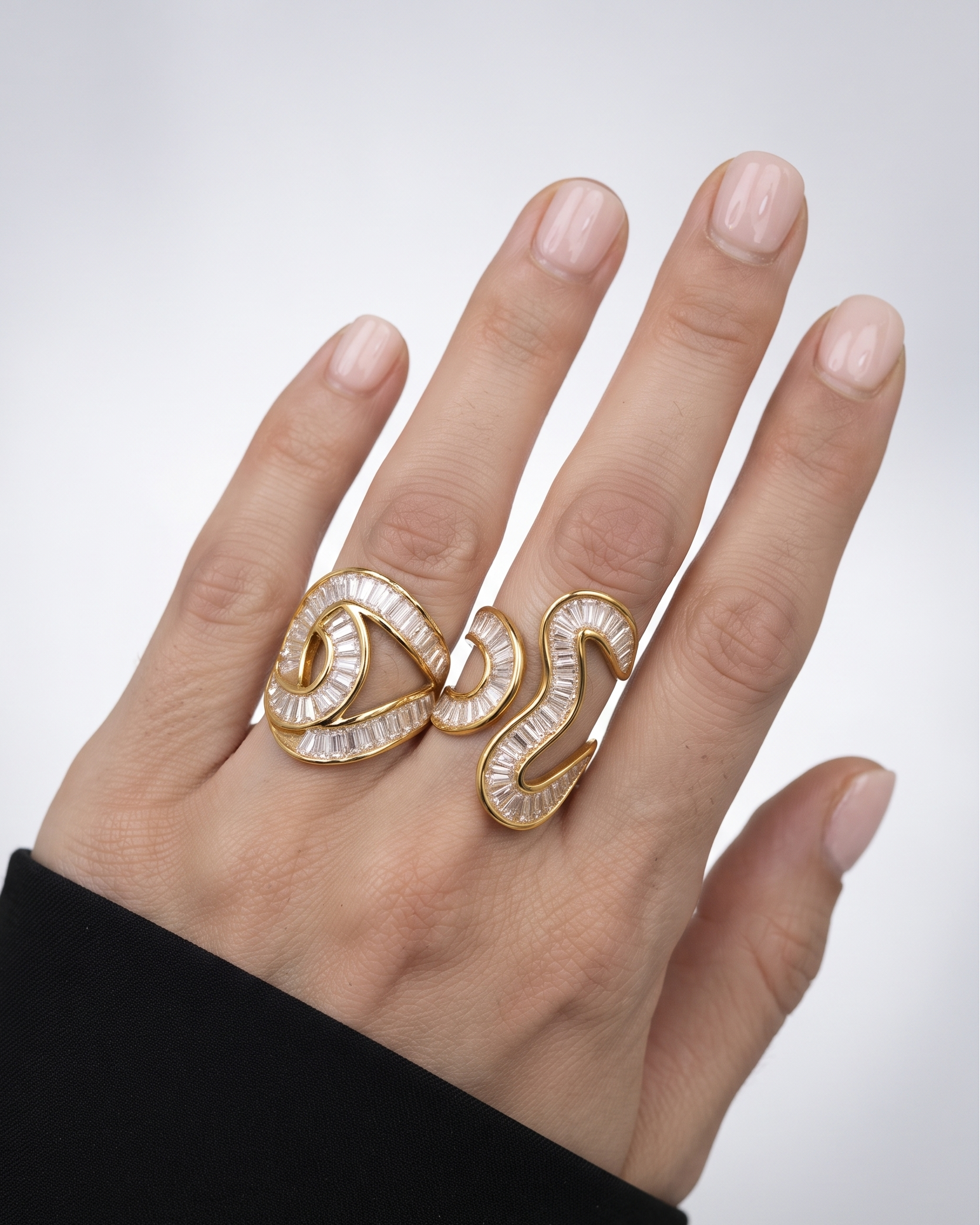 A close-up of a hand wearing a gold ring with two intertwined spiral motifs set with clear, faceted stones. The hand has a clean, neutral manicure and is wearing a black sleeve, set against a soft, light gray background.