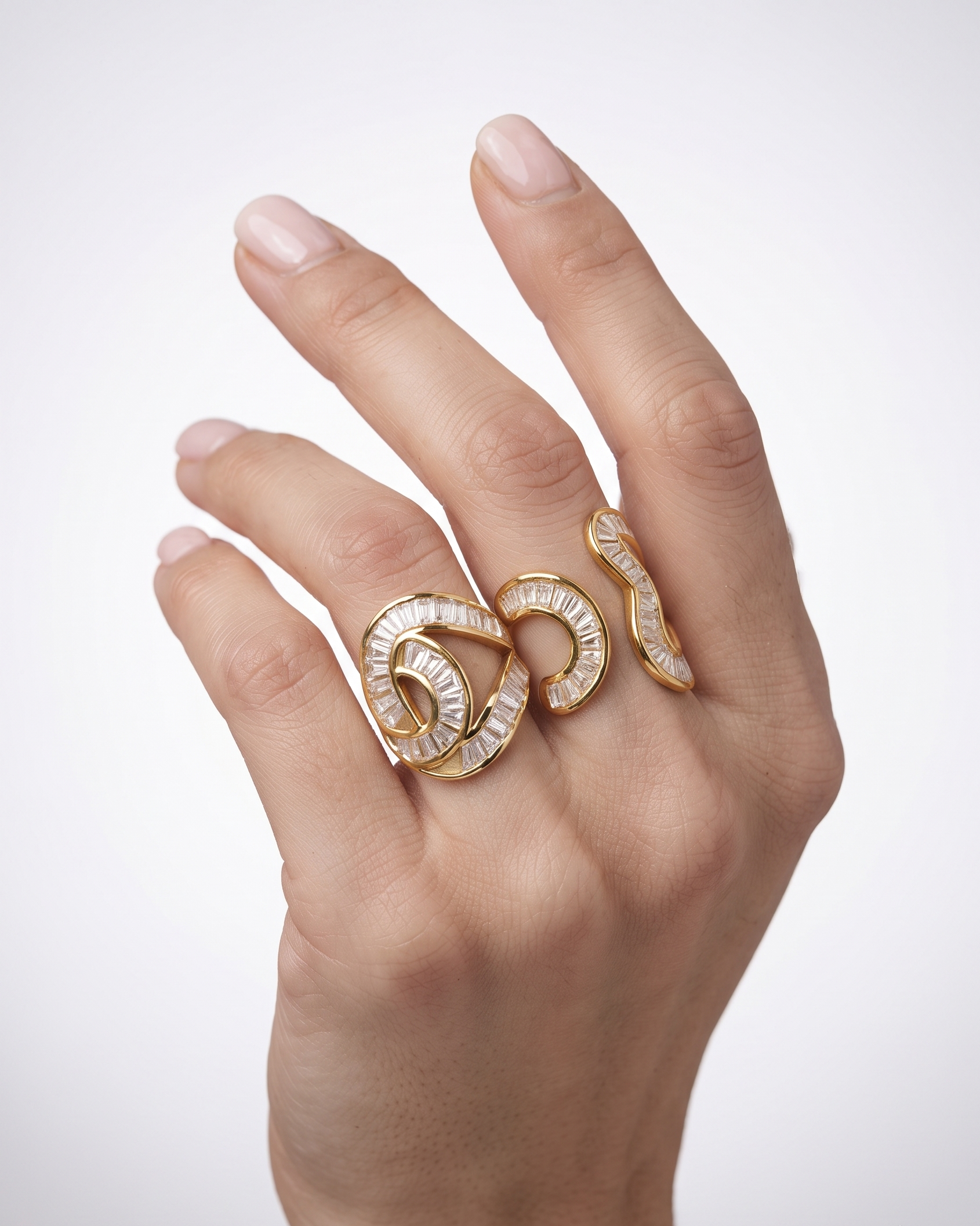 A close-up of a hand wearing two gold rings with textured, snake-like curved designs set with clear stones, against a light gray background.