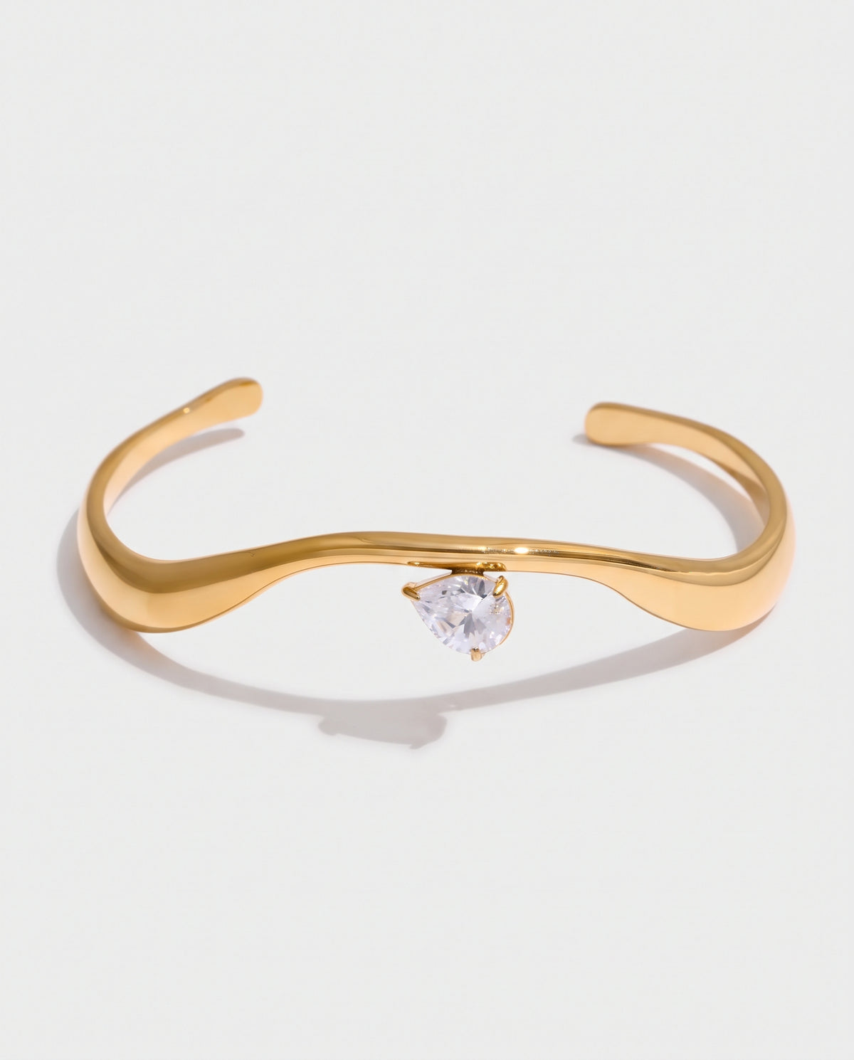 A gold-toned open cuff bracelet featuring a small, clear, heart-shaped gemstone at the center, displayed on a clean white background with a soft shadow.