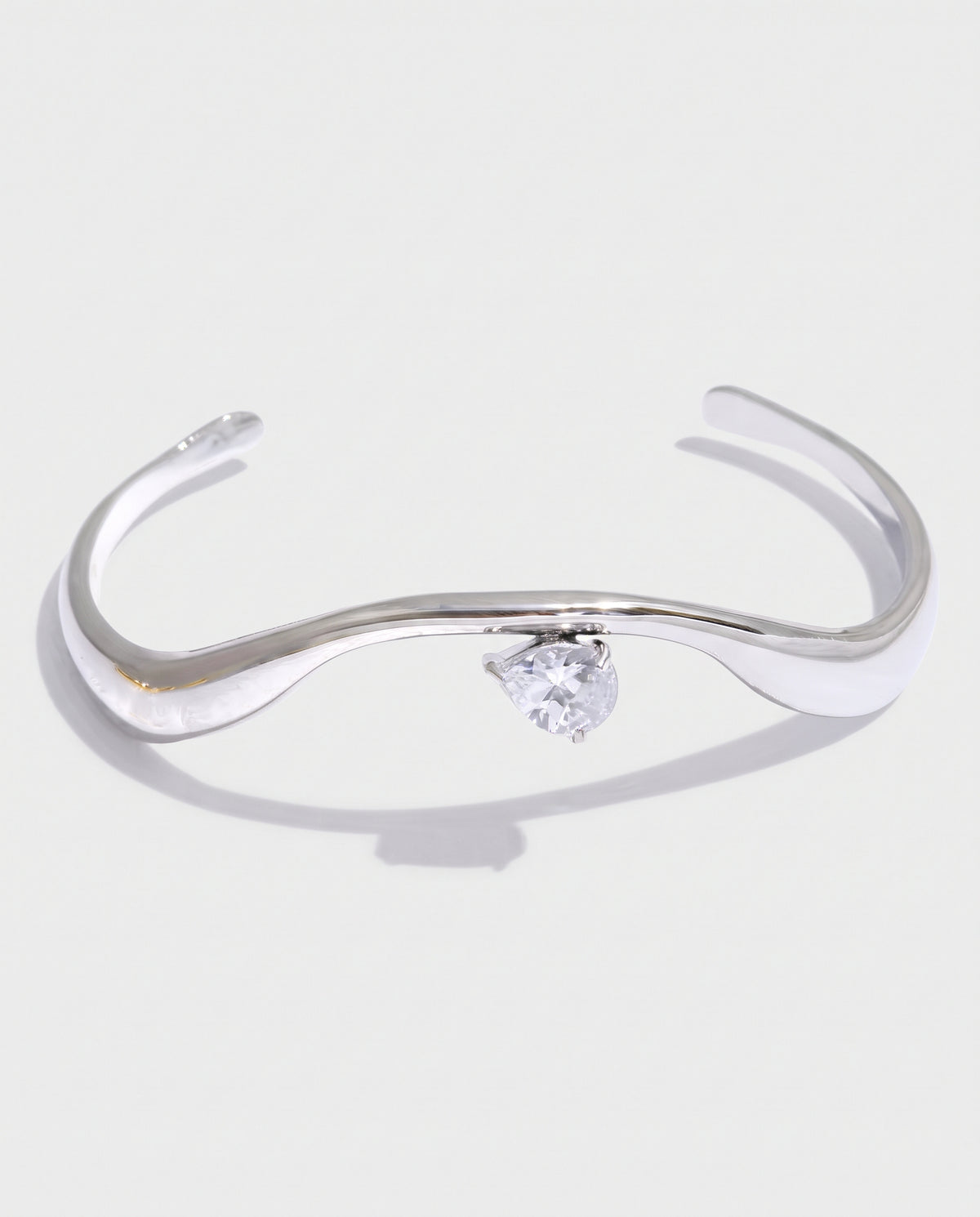 A polished silver-tone open cuff bracelet featuring a central triangular clear gemstone, set against a light gray background with a soft shadow.