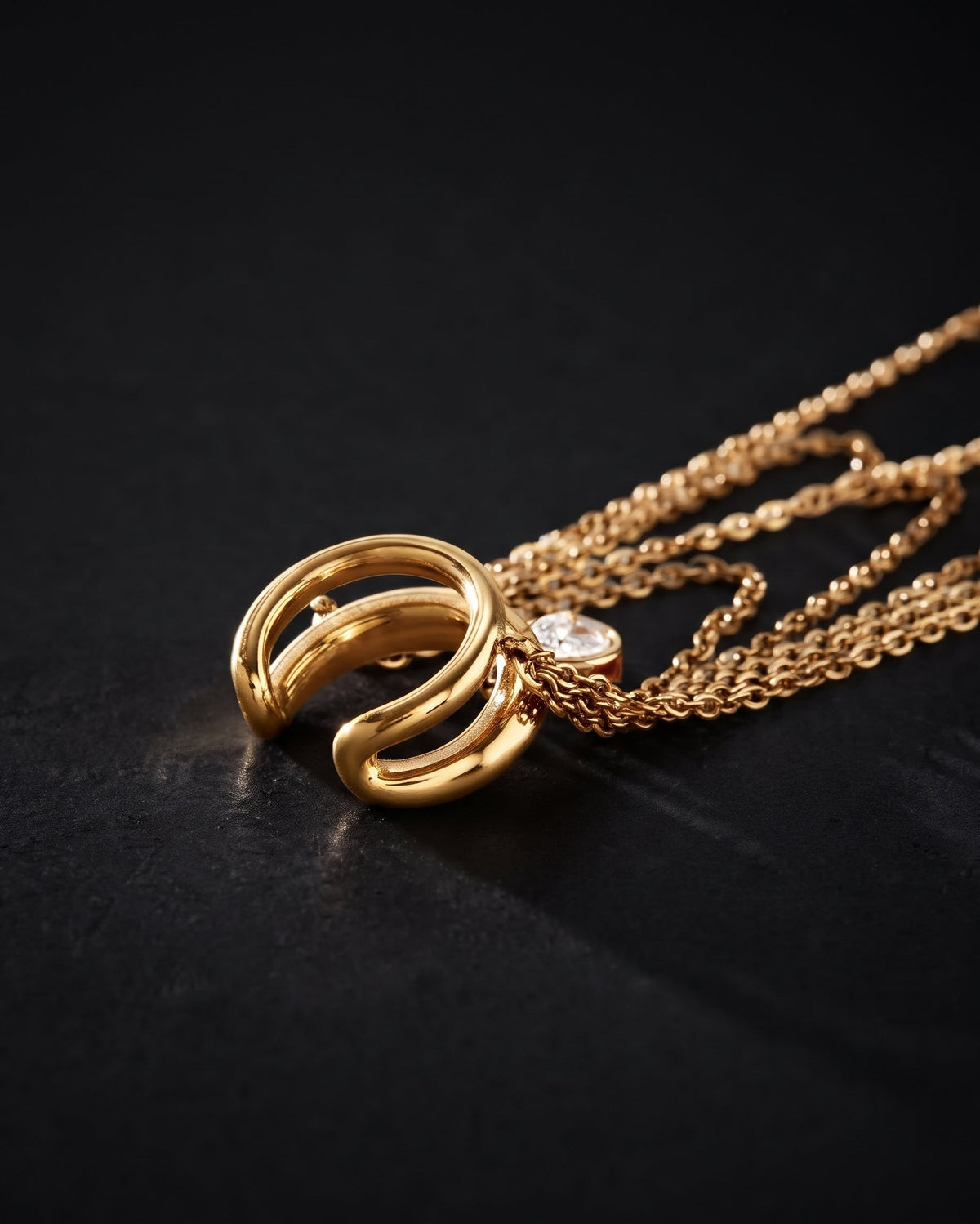 A gold pendant necklace featuring a sculptural, multi-looped open design with a small clear gemstone accent, resting on a dark textured surface.