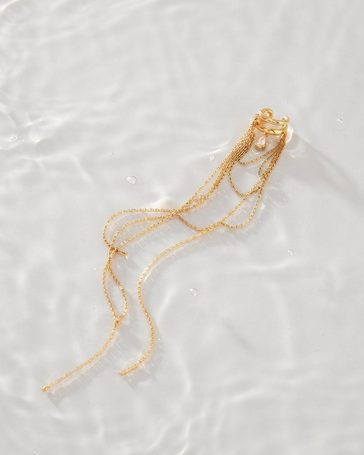 A delicate gold chain necklace with a small pearl pendant and multiple long trailing chains, photographed on a softly rippled white water surface.