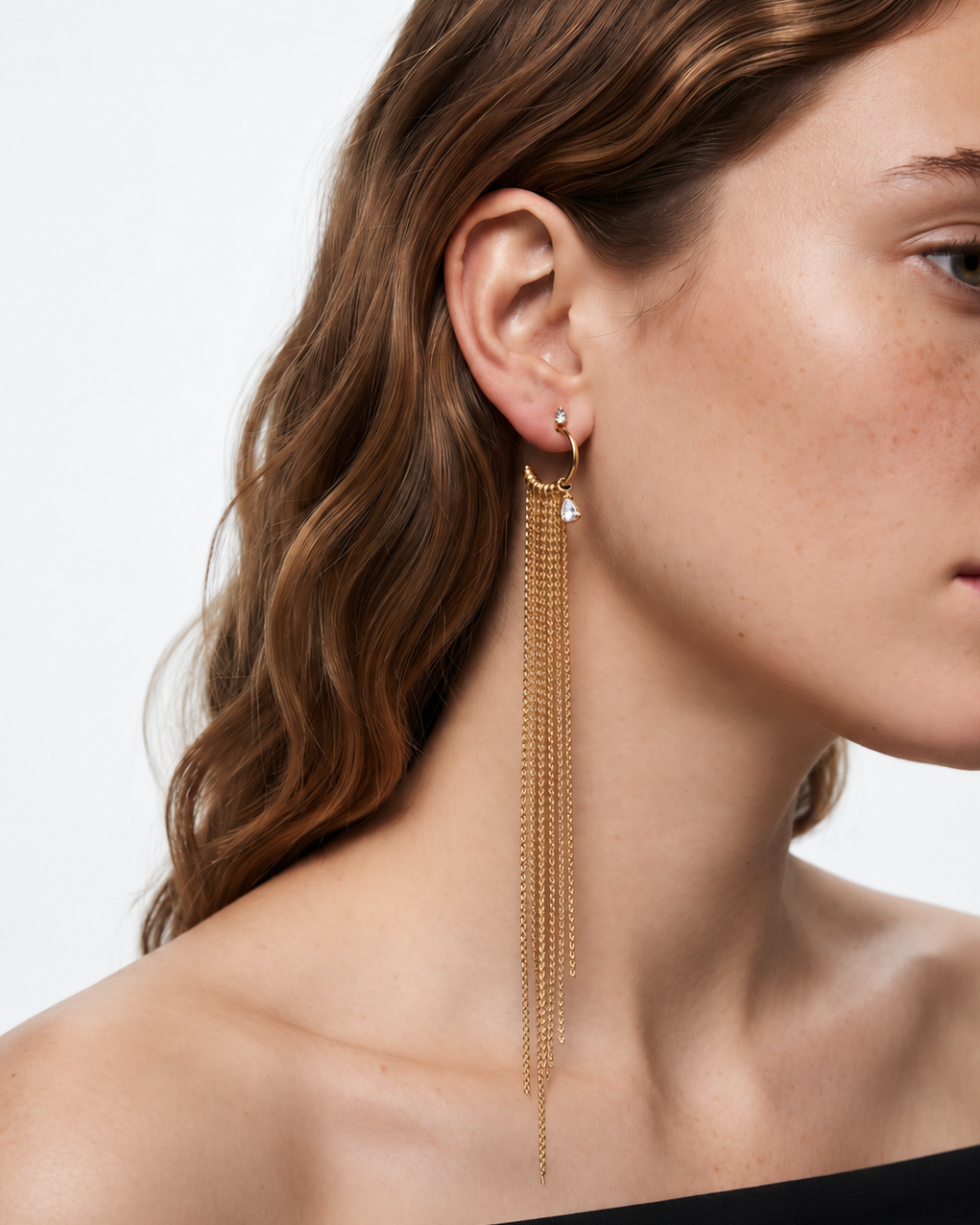 A close-up of a woman wearing a long, delicate gold dangle earring with multiple cascading chains and a small clear stone, shown against a plain light background.
