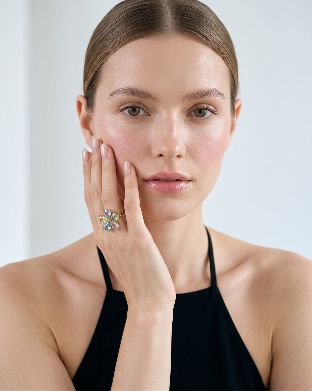 A close-up portrait of a woman with her hand resting against her cheek, wearing a colorful floral ring with pastel pink, blue, yellow, and green stones on a black halter top against a light gray background.