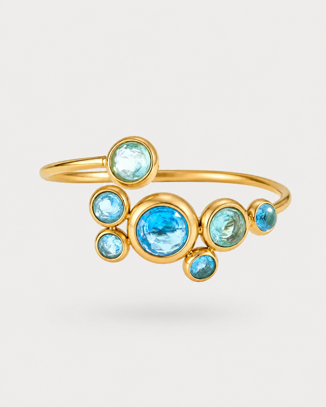 A gold-plated open cuff ring featuring a cluster of round-cut light blue gemstones in bezel settings, displayed on a clean light gray background.