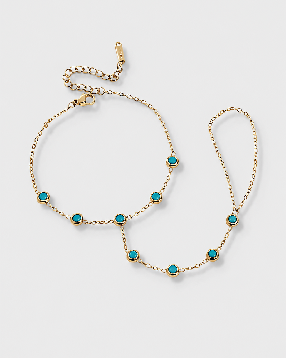 A gold chain anklet with seven small round turquoise-blue stones set in gold bezels, displayed on a light gray background.