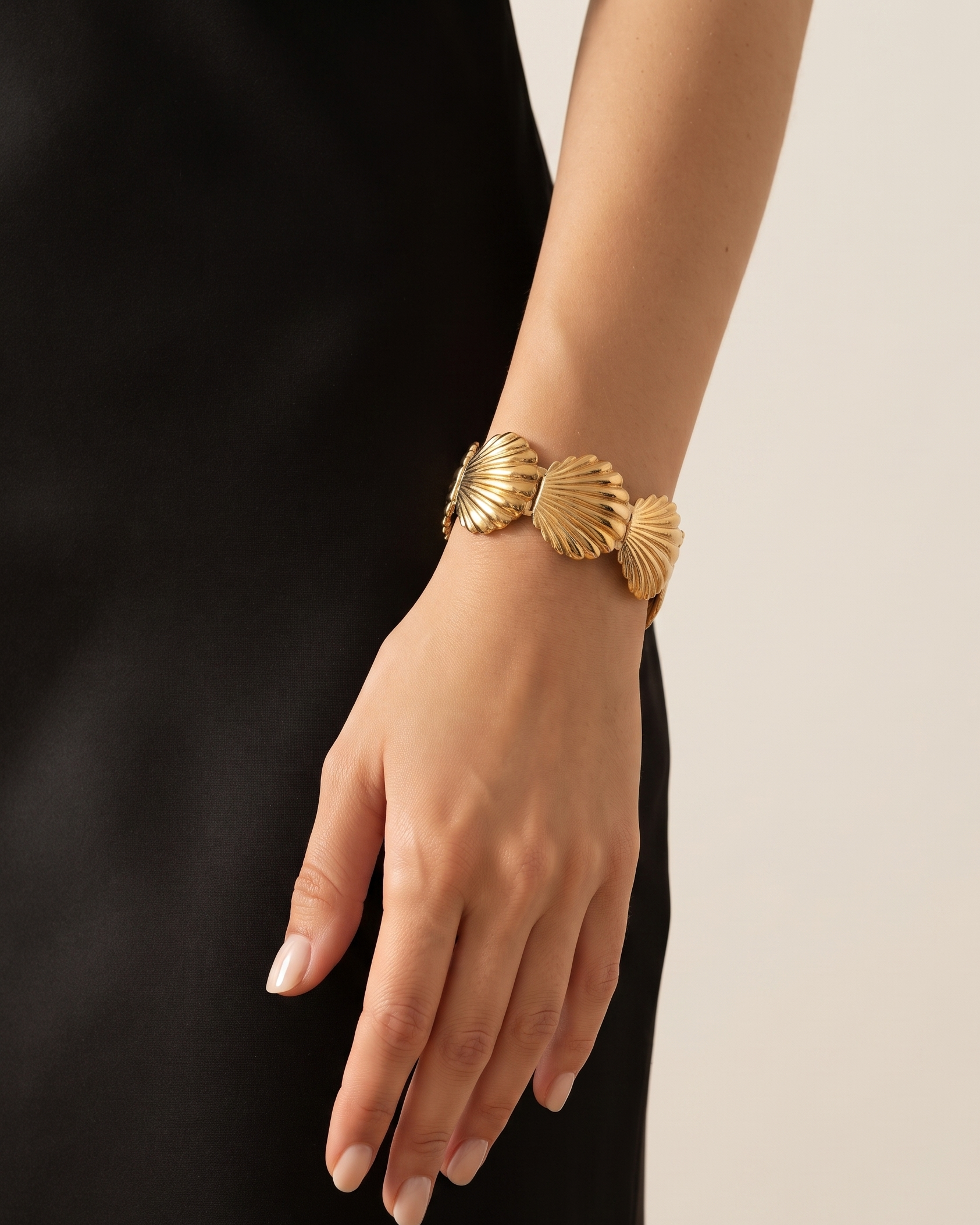 A close-up of a model's hand and wrist wearing a wide gold cuff bracelet with three textured shell-like links, styled with a black dress against a light beige background.
