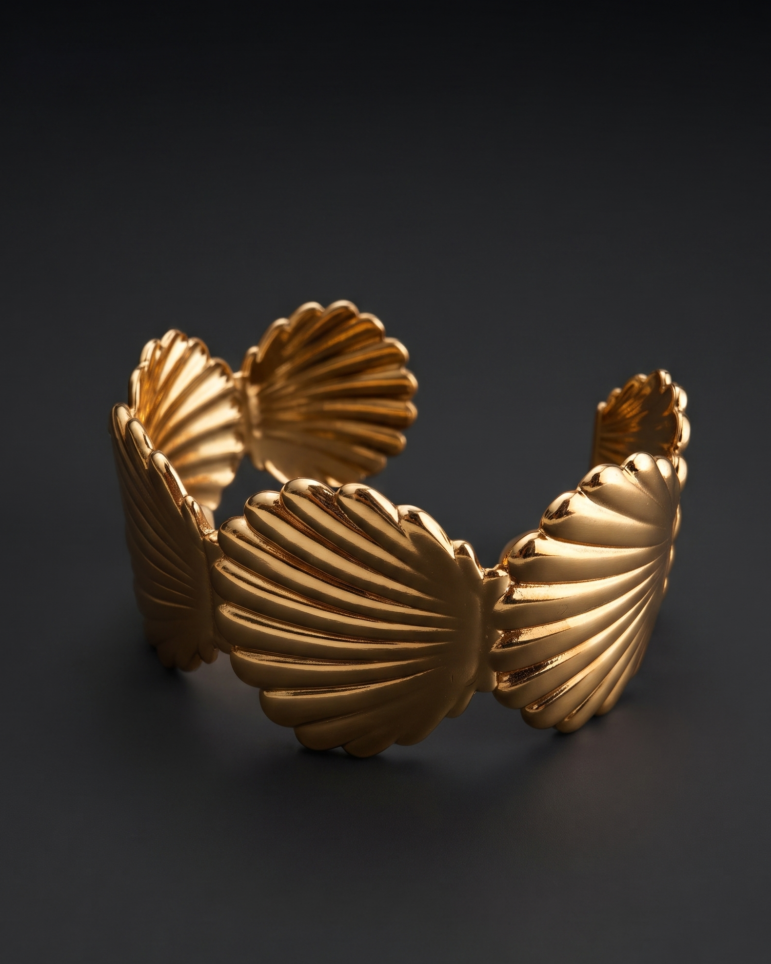 A gold-toned cuff bracelet with a sculptural, ribbed shell-like design, photographed on a dark gray background.
