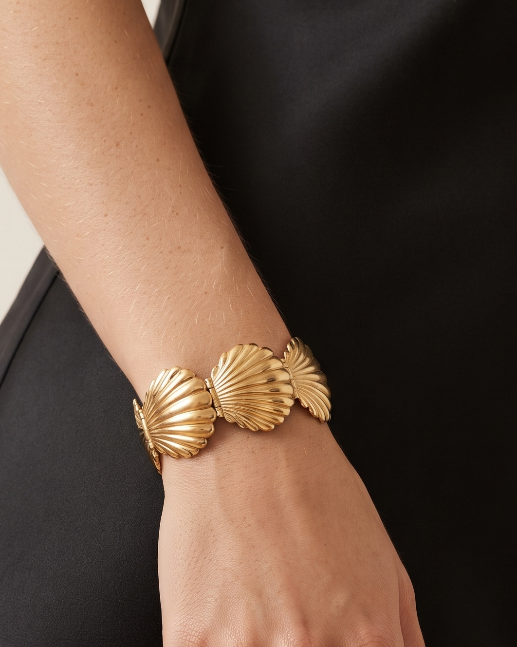 A close-up of a person's hand and forearm wearing a wide gold cuff bracelet with three shell-like segments, against a black dress and light beige background.