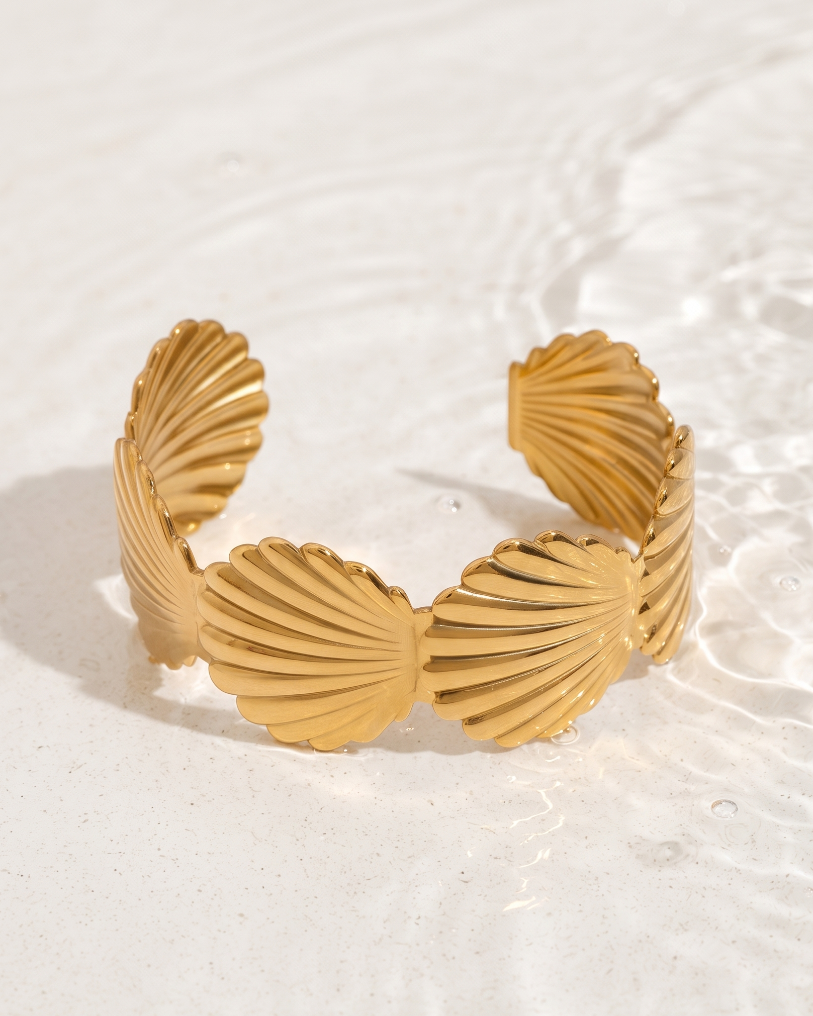A gold-toned cuff bracelet with a textured, ribbed scallop or shell-like design, resting on a white sandy surface with water ripples in the background.