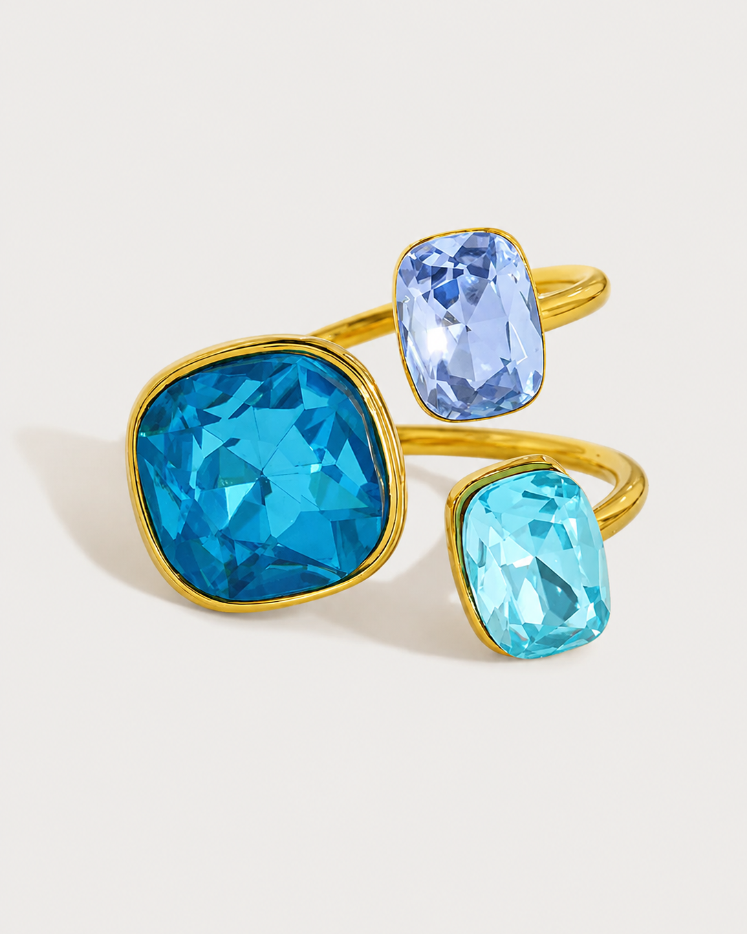 A gold-tone double ring set featuring three faceted gemstones in shades of blue, including a large bright blue cushion-cut stone, a medium periwinkle blue oval stone, and a light aqua blue square stone, photographed on a clean off-white background.