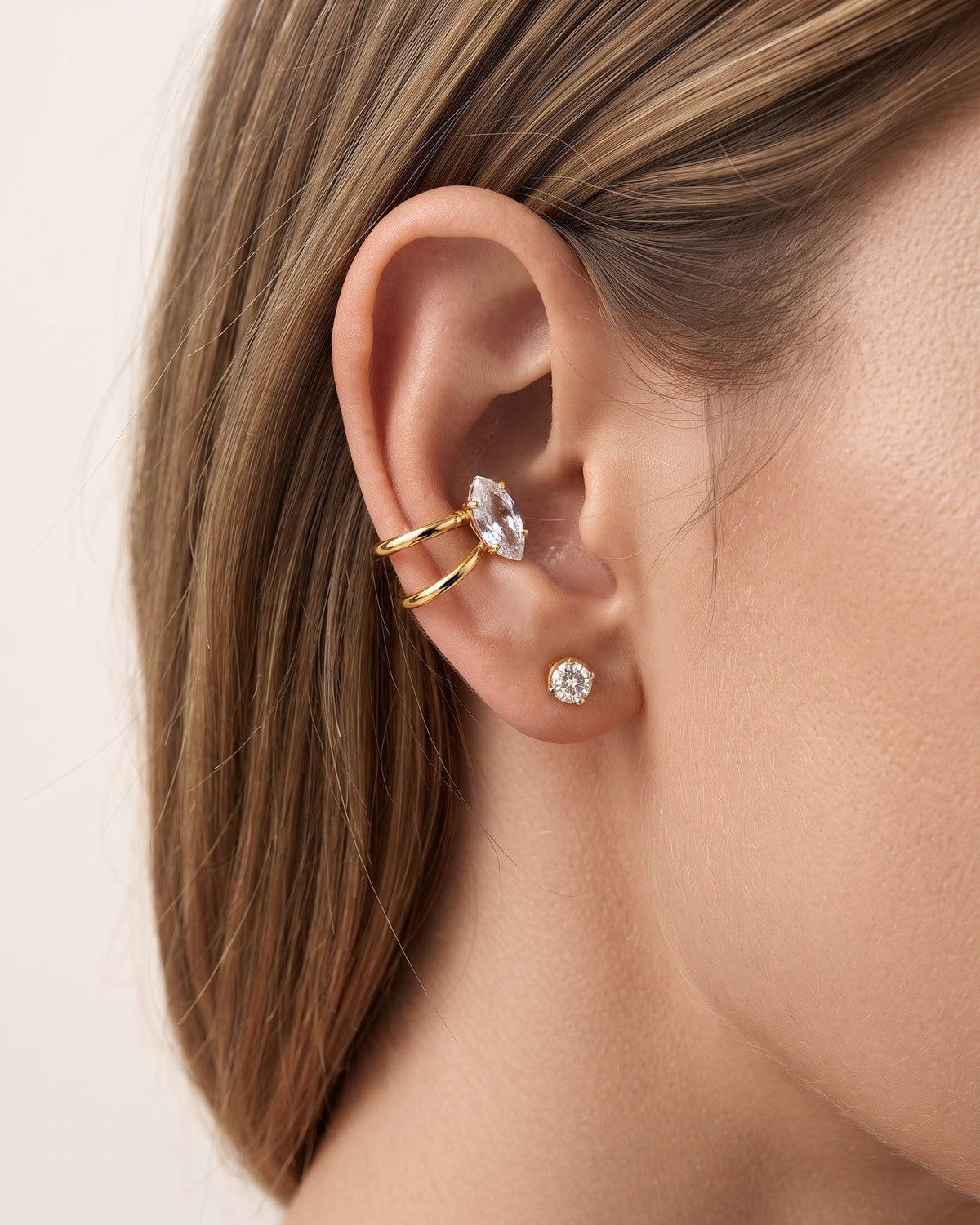 Close-up of a model's ear wearing a gold ear cuff with a marquise-cut clear gemstone and a small gold stud earring with a round clear gemstone.
