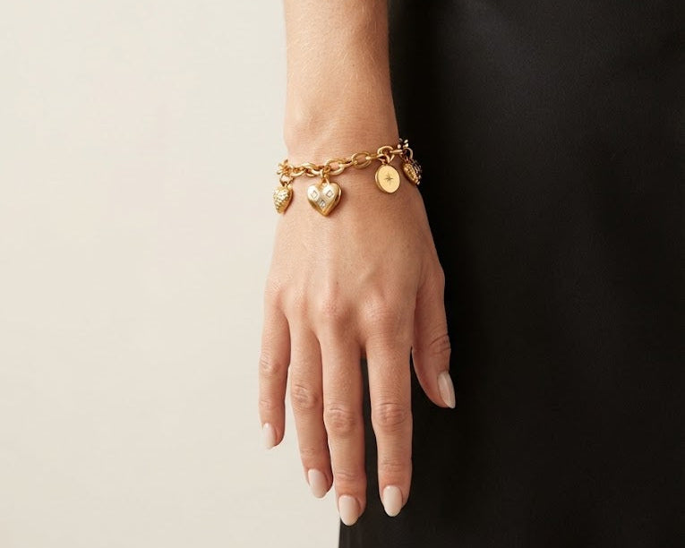 A gold charm bracelet with heart and disc charms worn on a model's wrist, shown against a neutral light beige background and a black dress.