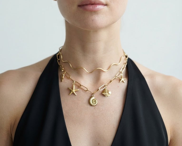 A blonde woman wearing a layered gold necklace with starfish, shell, and sun pendant charms, paired with a black halter top against a light gray background.