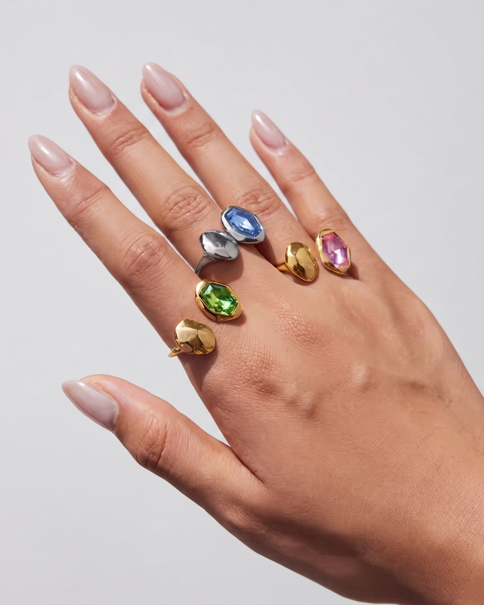 A hand with manicured nails wearing six gold rings, each featuring a different colored gemstone (blue, silver, pink, green, and yellow), set against a plain light gray background.