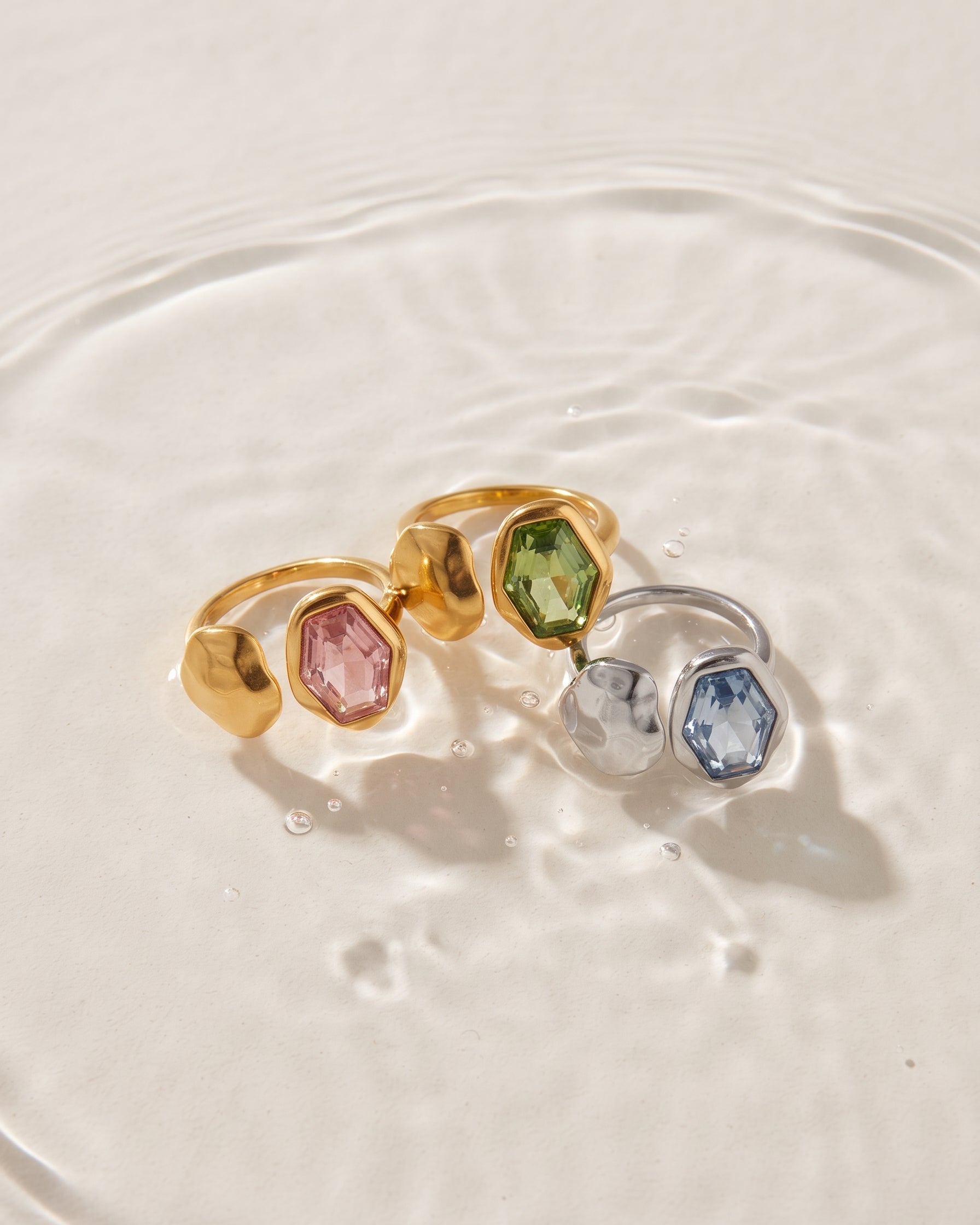 Three textured metal rings with faceted gemstone accents are arranged in shallow water. The rings feature gold, silver, and rose gold bands with pink, green, clear, and blue stones.