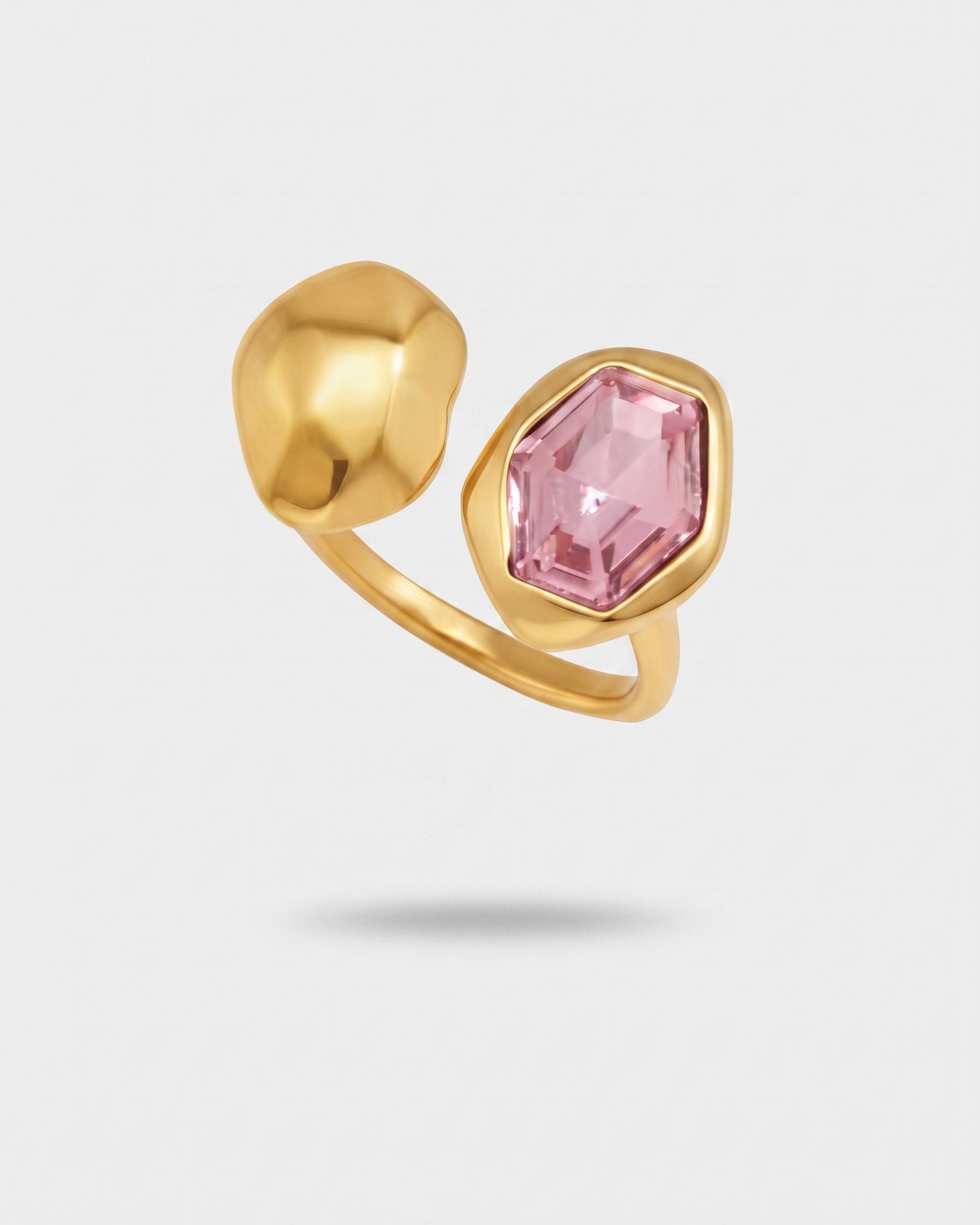 A gold ring featuring a faceted pink gemstone and a sculptural gold bead, shown on a light gray background.