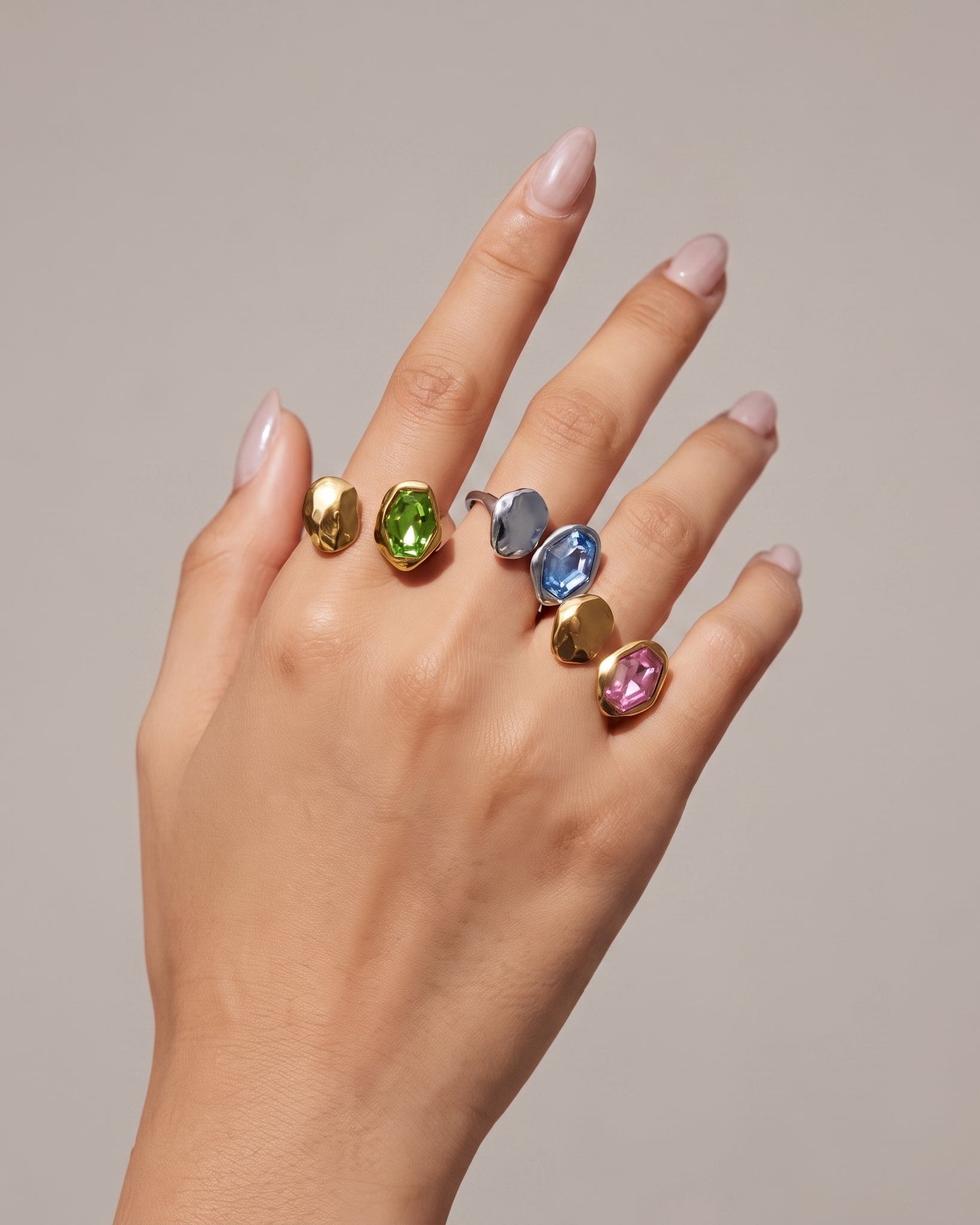 A hand with light pink manicured nails wearing five colorful rings, including gold, green, blue, pink, and silver oval gemstone rings, against a solid beige background.