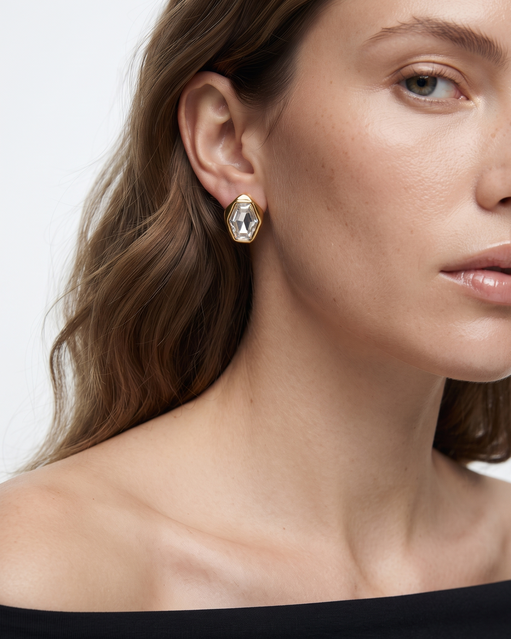 A close-up of a model wearing a gold geometric stud earring with a clear faceted gemstone. The shot focuses on the earring and the model's ear, neck, and shoulder against a clean white background.