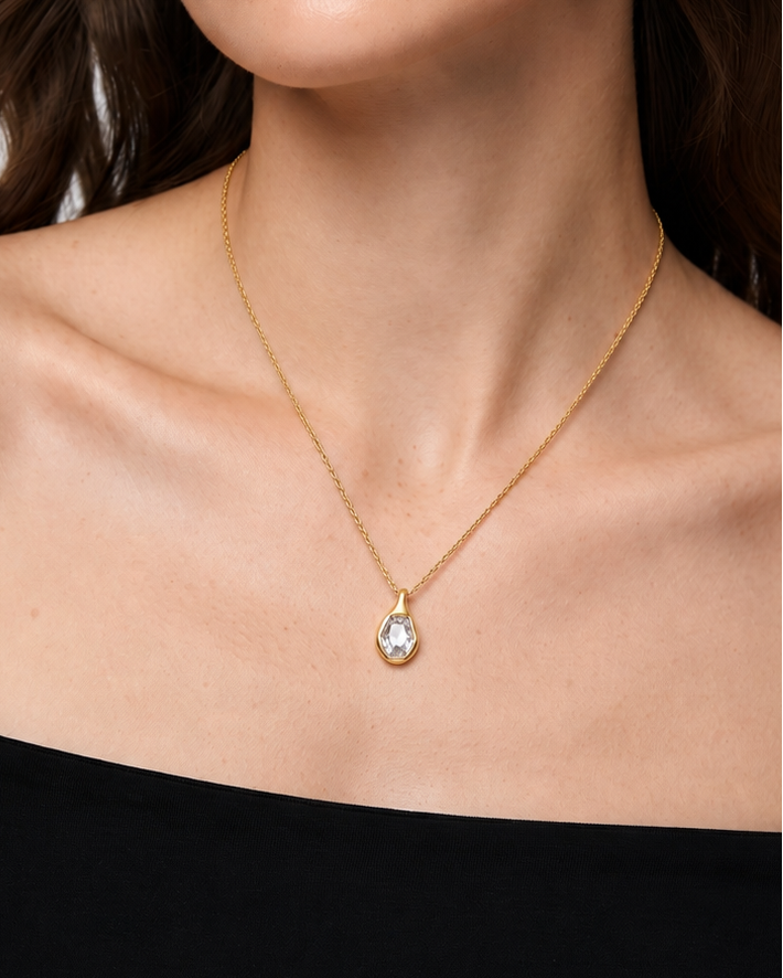 A close-up of a woman wearing a delicate gold chain necklace with a teardrop-shaped clear gemstone pendant, styled with a black off-the-shoulder top against a light gray background.