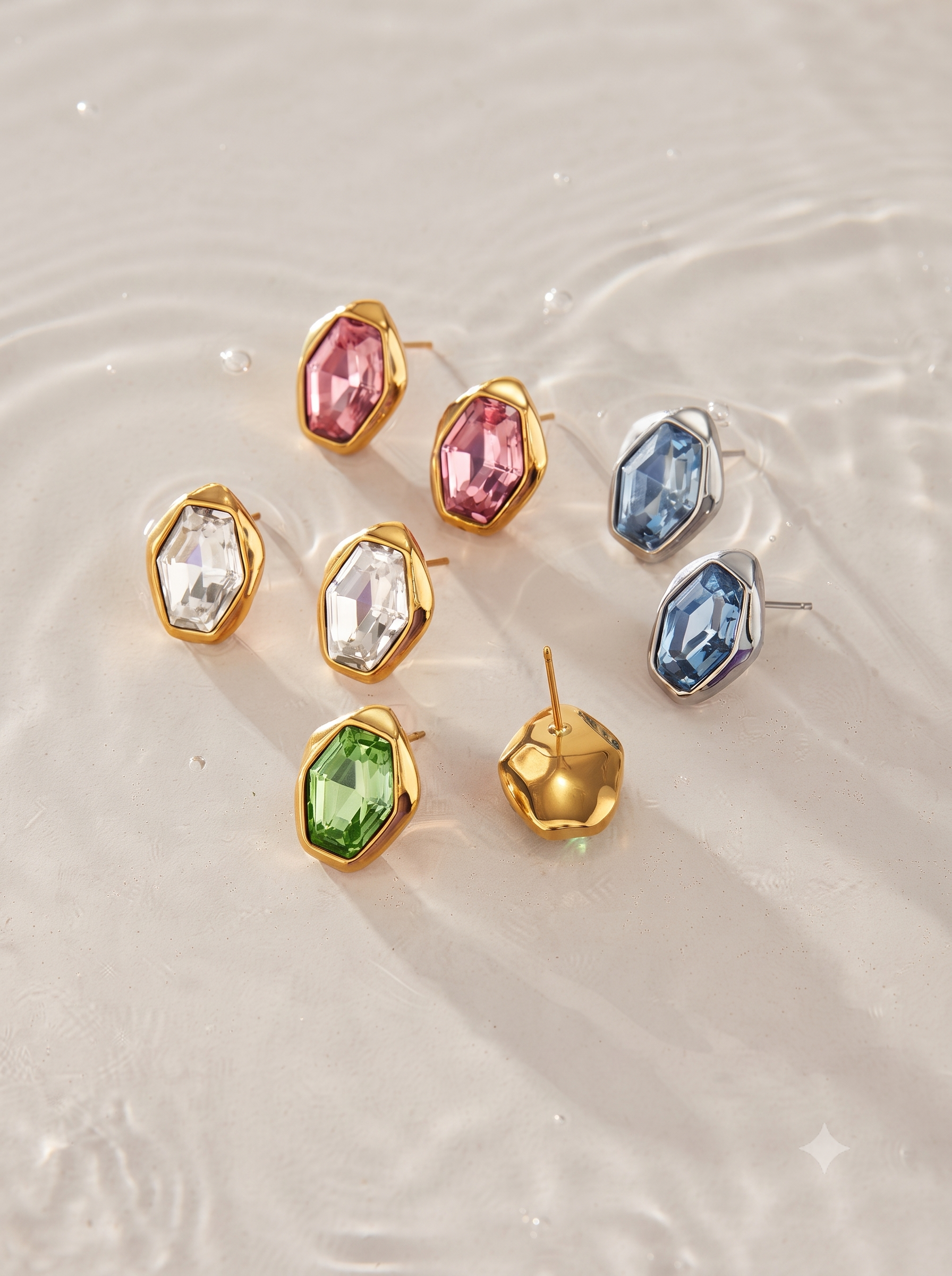 A set of colorful geometric stud earrings arranged on a light beige textured surface with soft water ripples. The earrings feature faceted gemstone-like tops in pink, blue, green, and clear crystal, set in gold and silver-tone metal.