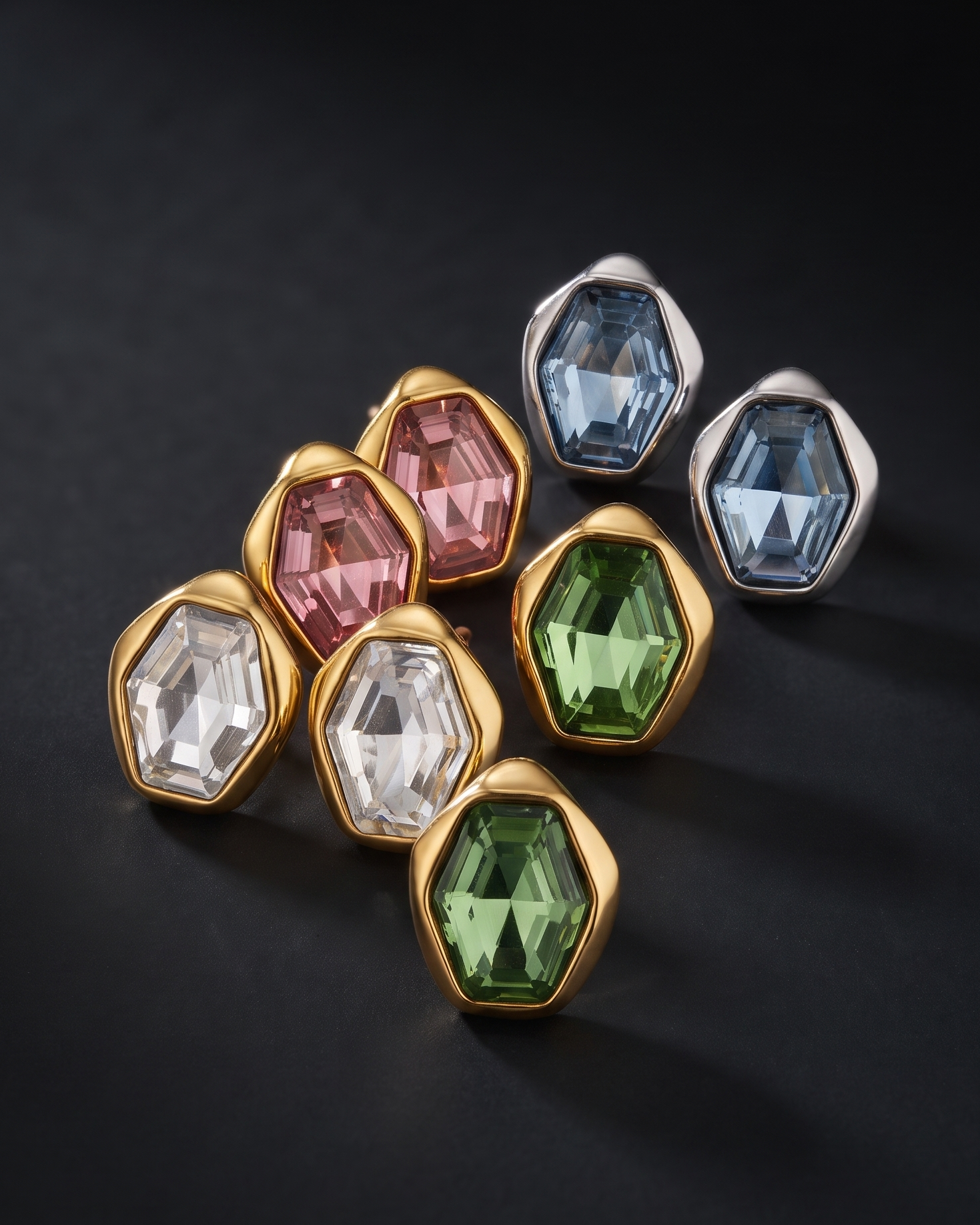 A set of faceted gemstone stud earrings in gold and silver bezel settings, arranged on a dark black background. The earrings feature green, pink, blue, clear, and light blue stones.