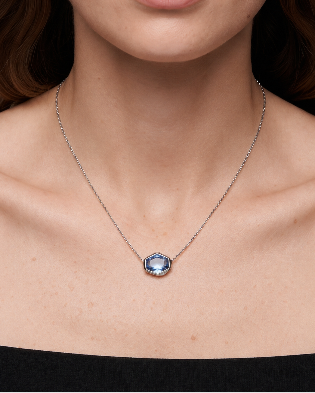 A close-up of a model wearing a delicate silver chain necklace with a small hexagonal pendant featuring a light blue faceted gemstone.