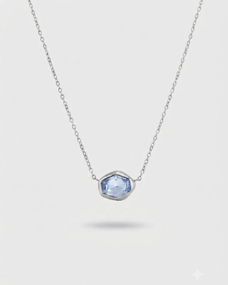 A delicate silver chain necklace featuring a small, irregular-cut light blue gemstone pendant in a silver bezel setting, shown against a light gray background.
