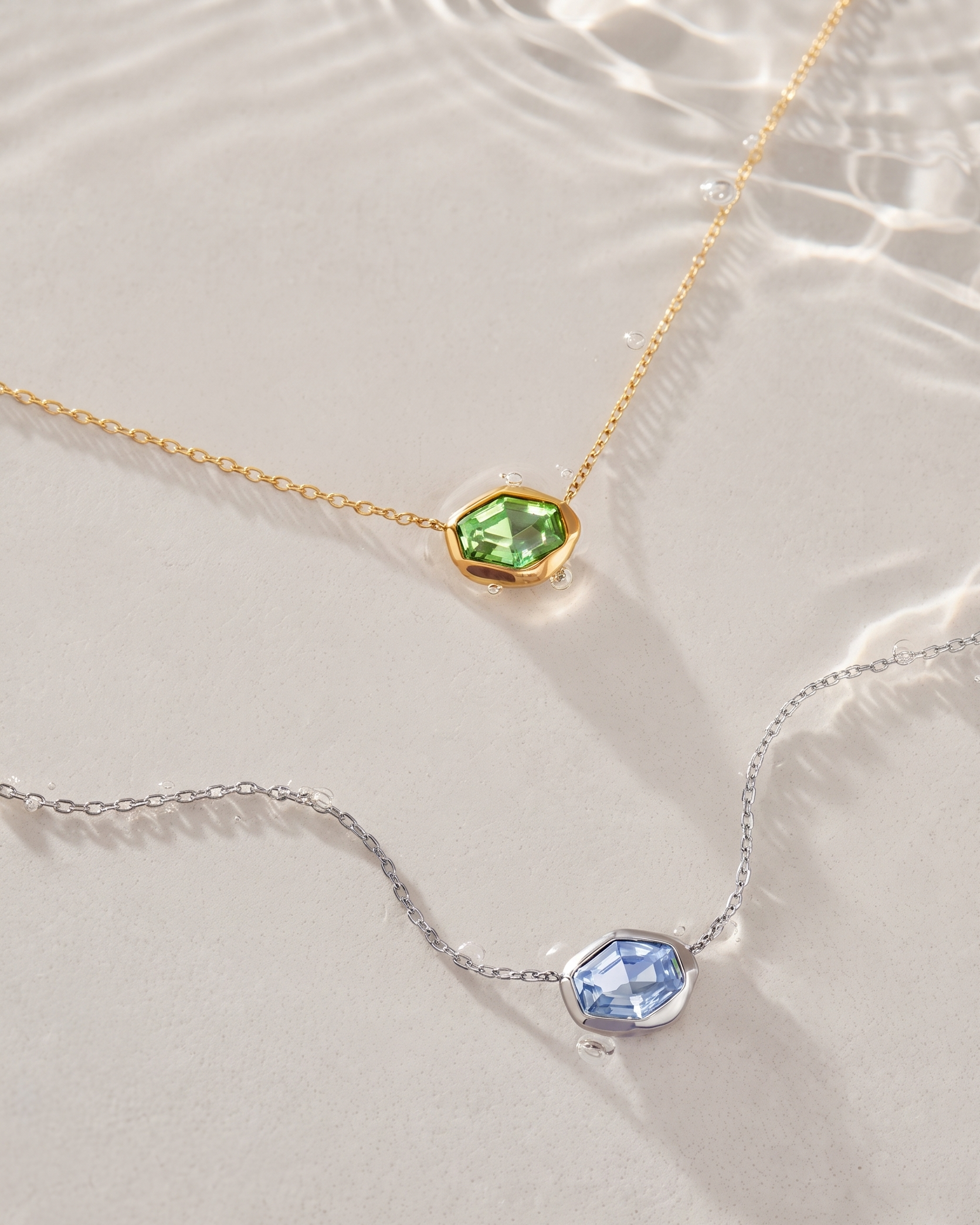 Two delicate necklace chains laid on a light beige surface with soft water-like ripples. One chain is gold with a faceted green gemstone pendant, and the other is silver with a faceted light blue gemstone pendant.
